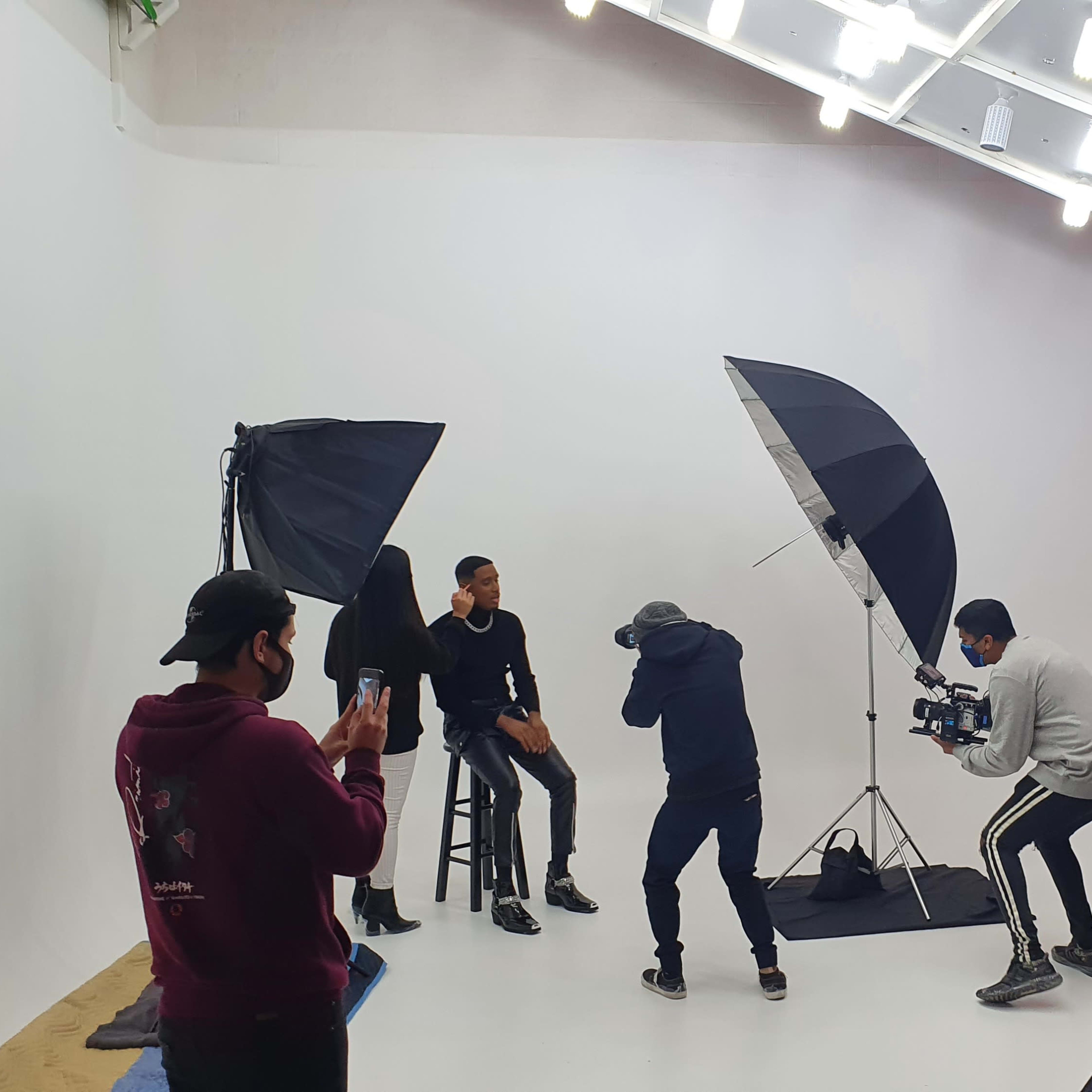 A group of people is filming a photoshoot in a studio with large softbox lights and a plain white background.