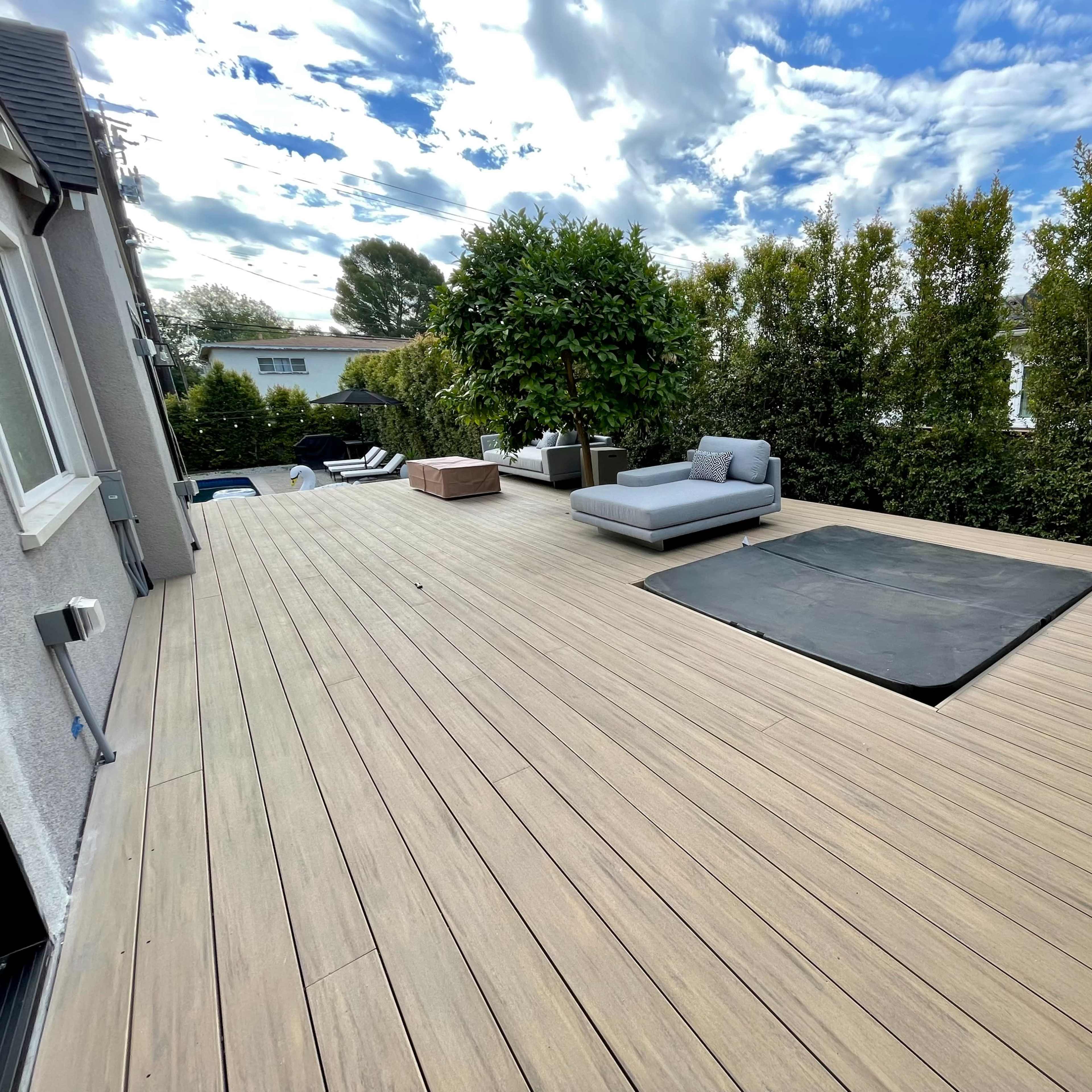 A spacious wooden deck features a sunken area next to a gray lounge chair and surrounding greenery.