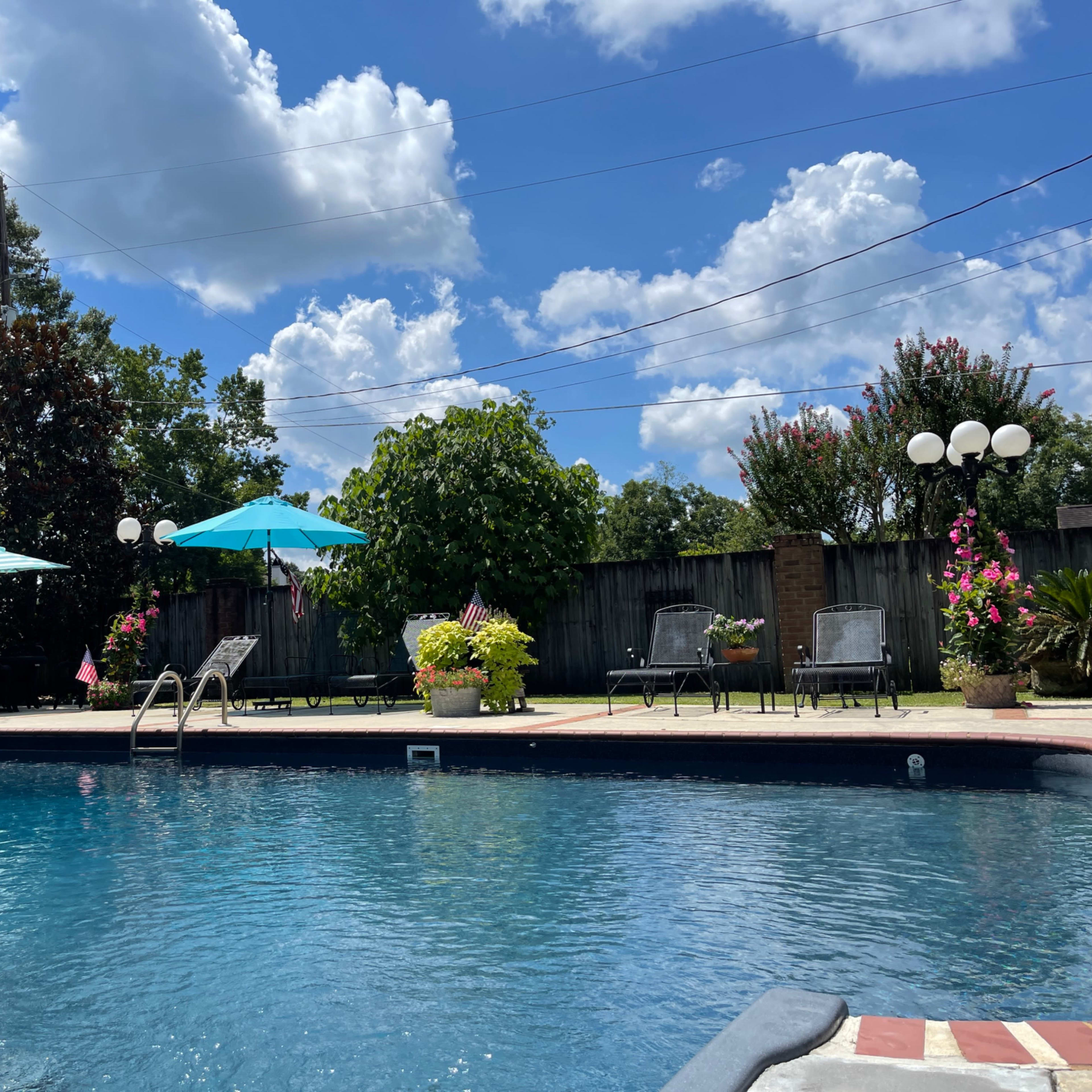 A clear pool surrounded by lounge chairs, umbrellas, and landscaped greenery under a partly cloudy sky.