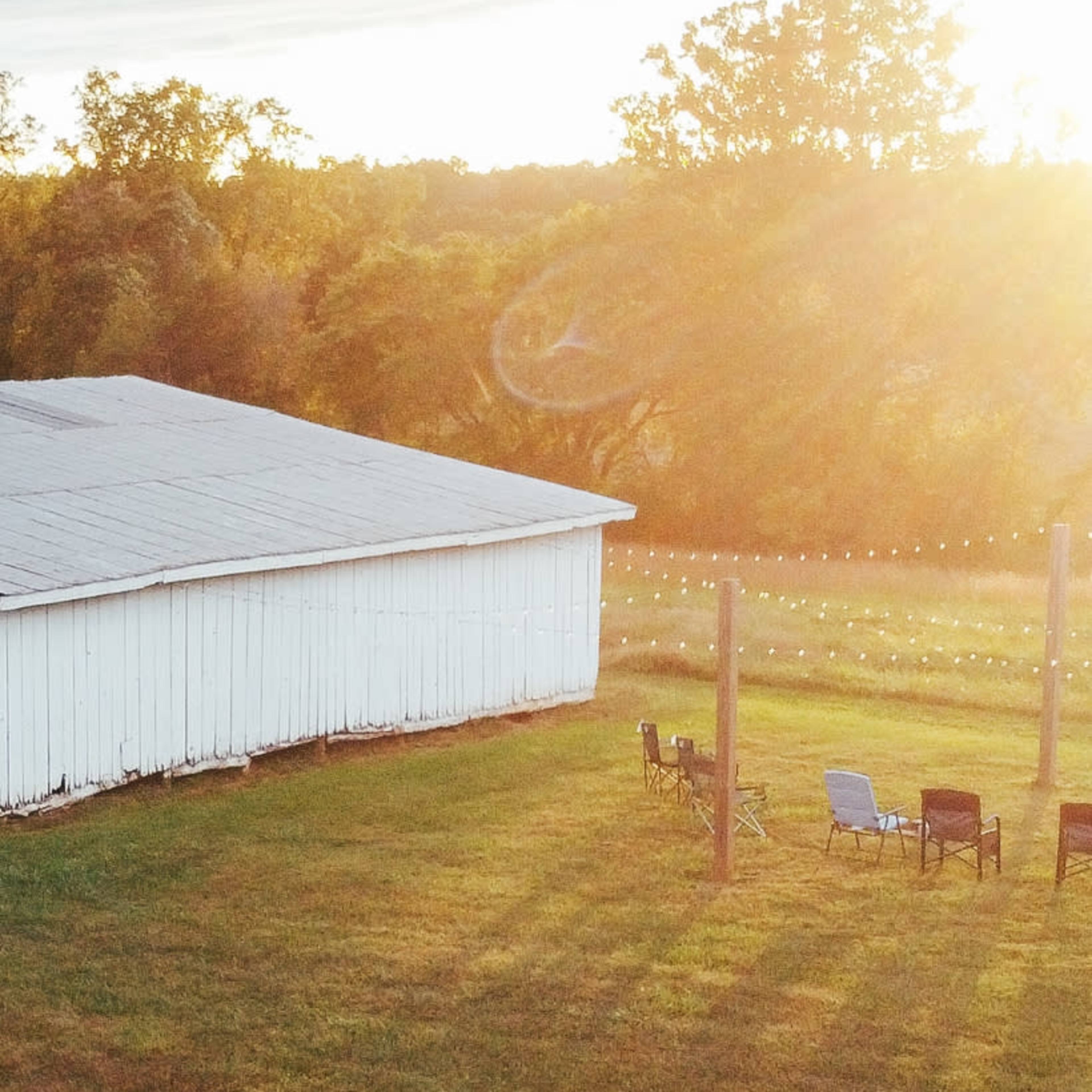 A white barn sits beside a green lawn illuminated by the setting sun, with a row of chairs and string lights in the background.