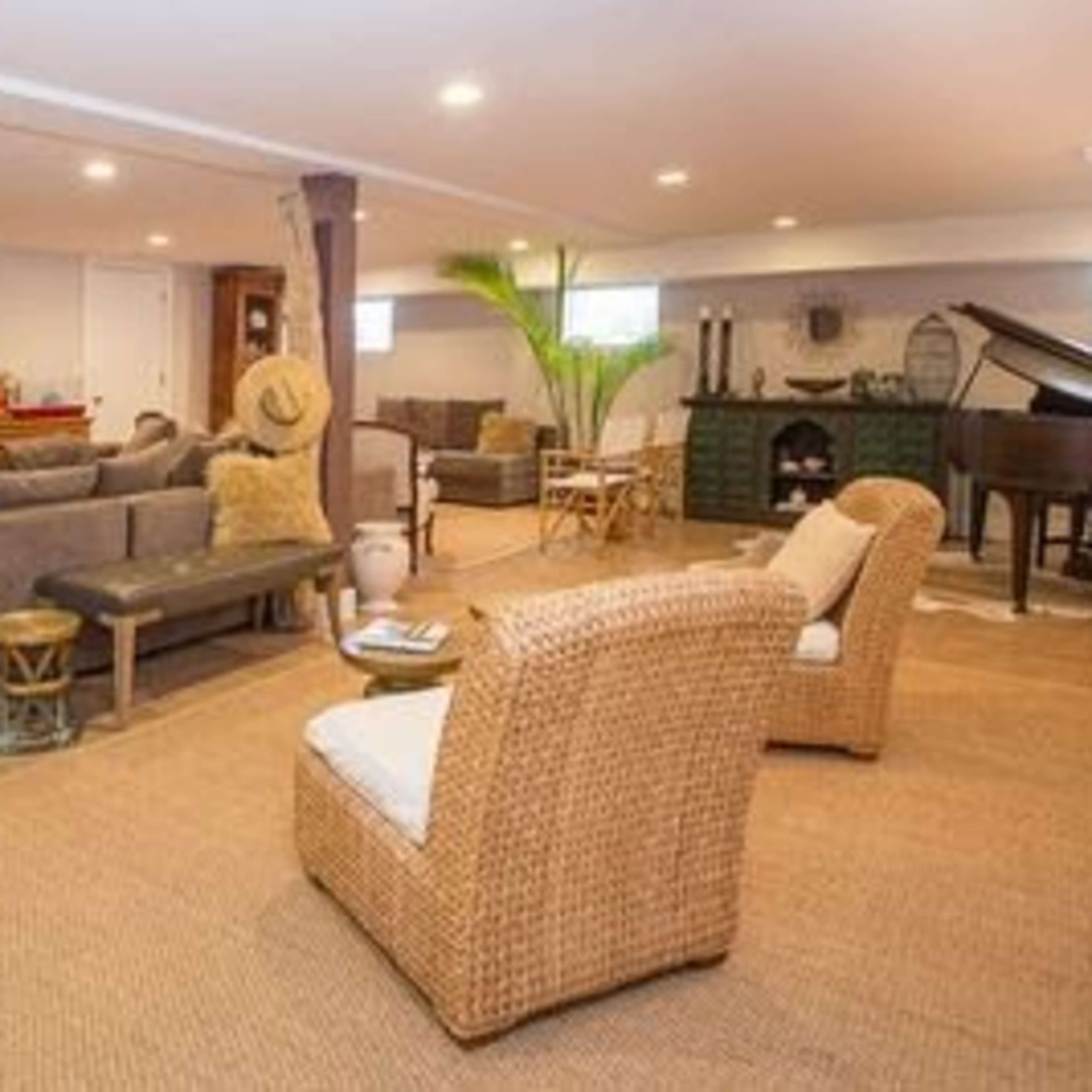 A spacious basement living area furnished with a sectional sofa, armchairs, a piano, a television, and decorative elements, featuring a warm color palette and natural light.