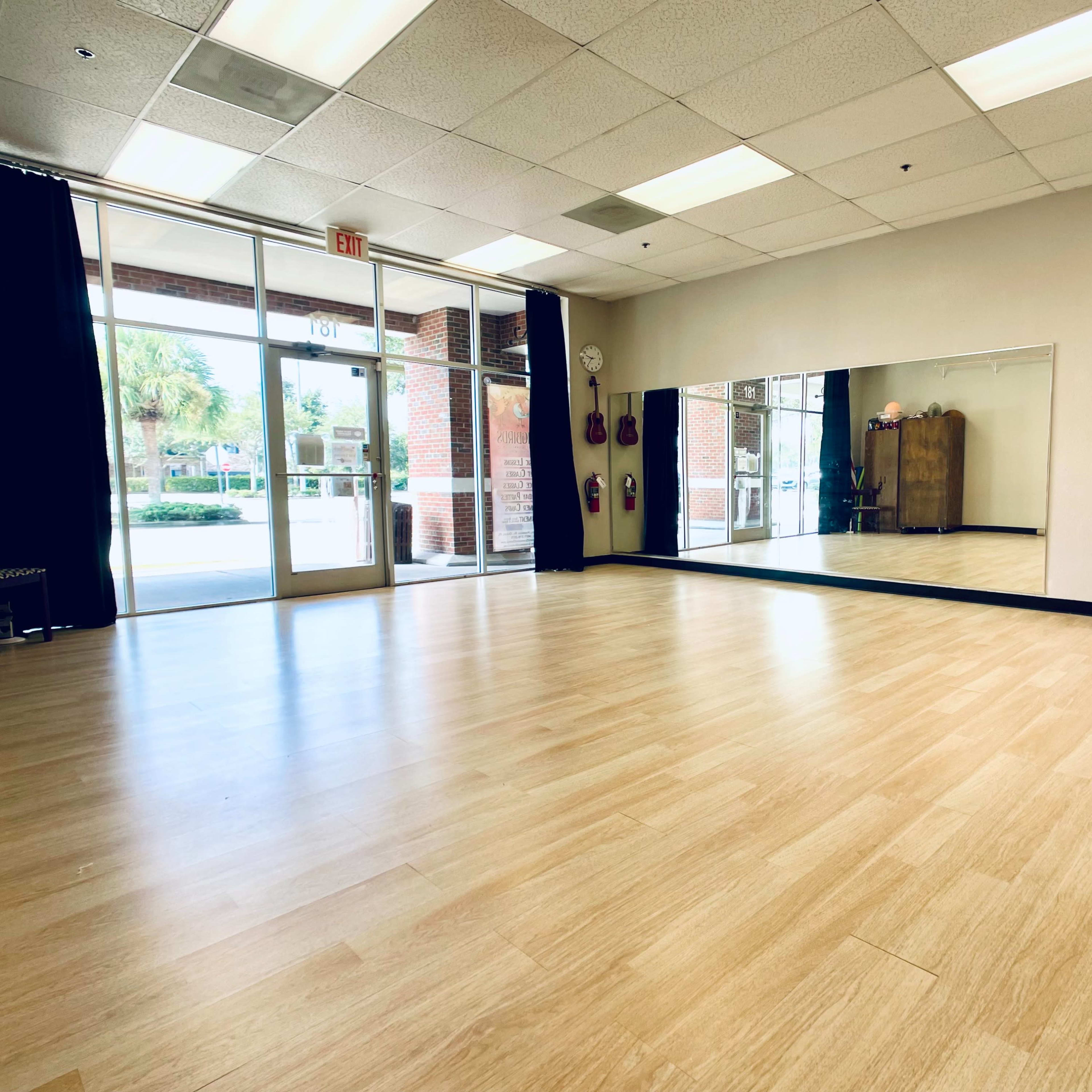 A spacious dance studio with wooden flooring, large mirrors on the wall, and natural light coming through the glass front doors.