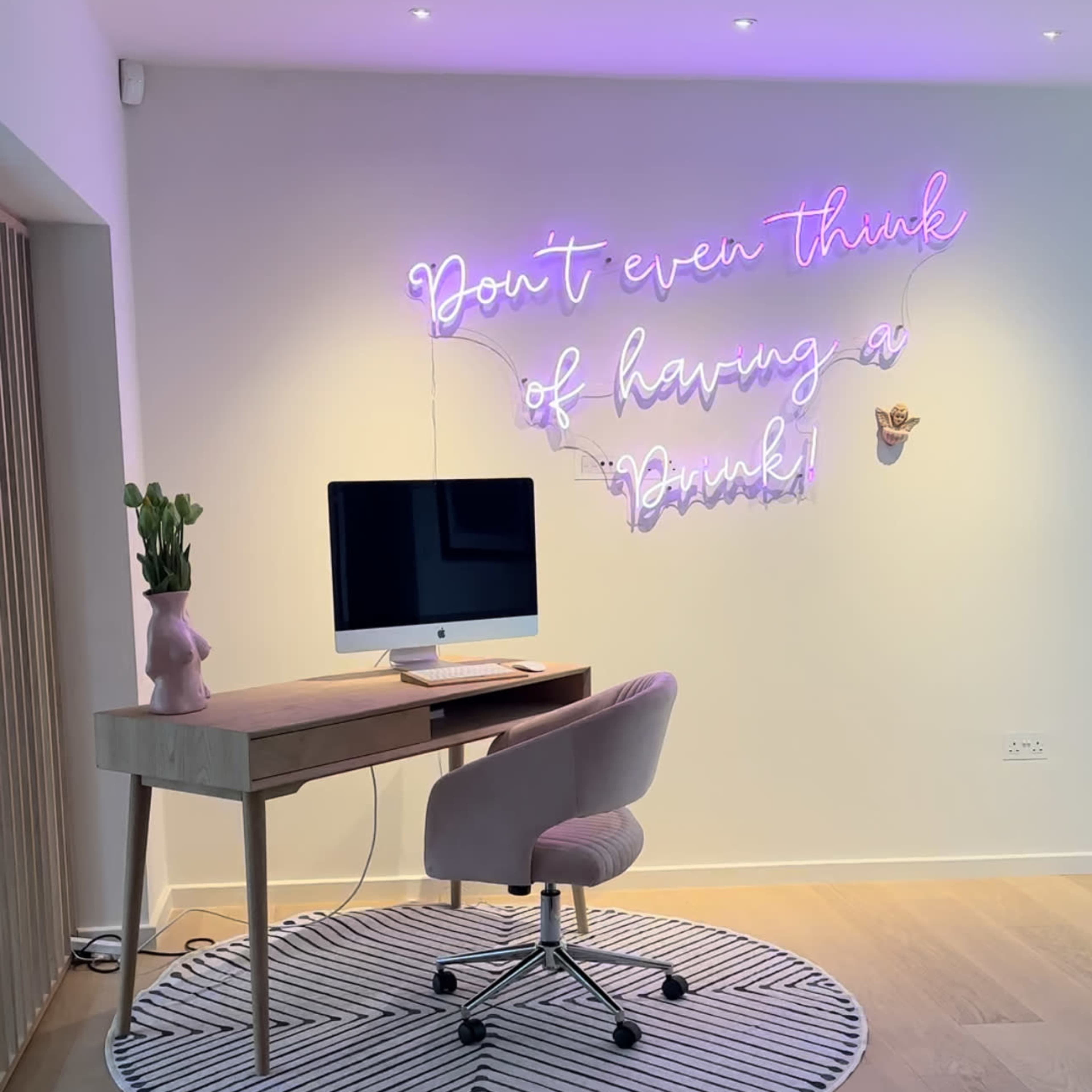 A modern workspace features a wooden desk with an iMac, a pink chair, and a neon sign on the wall that reads, "Don't even think of having a drink!"