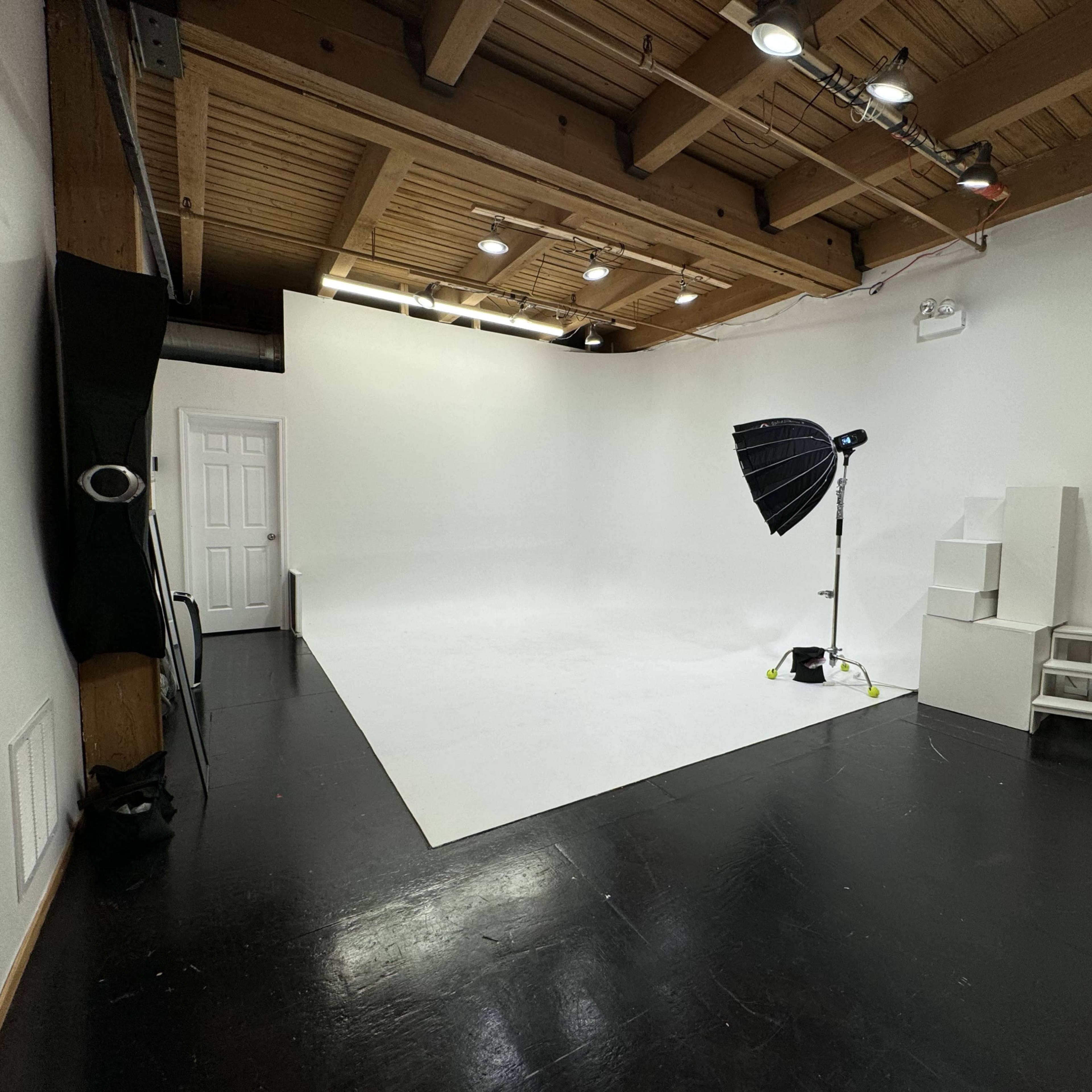 A spacious photography studio with a white backdrop, lighting equipment, and a few steps and a stool positioned in the corner.
