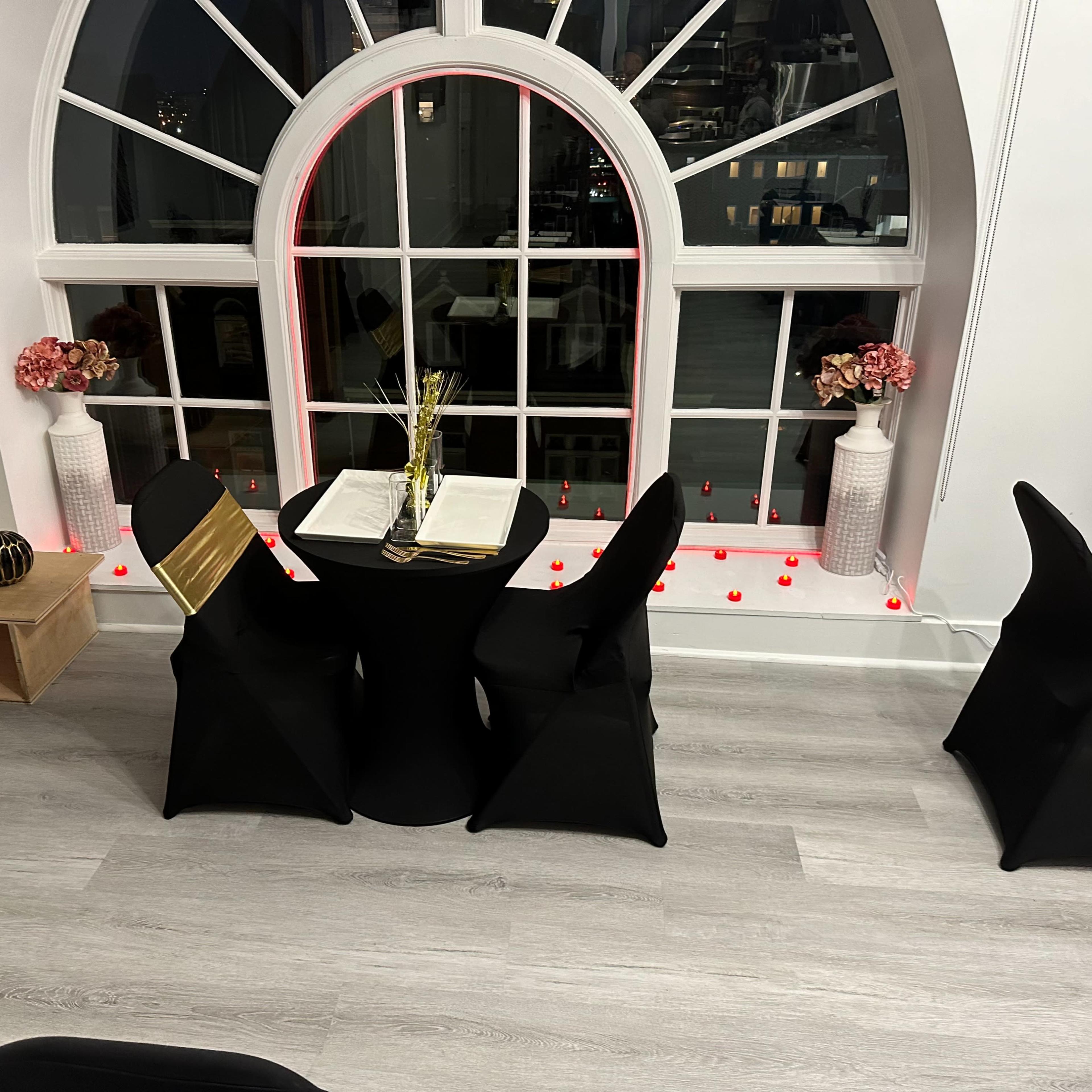 A small round table set for two near a large window with decorative vases and candles along the sill.