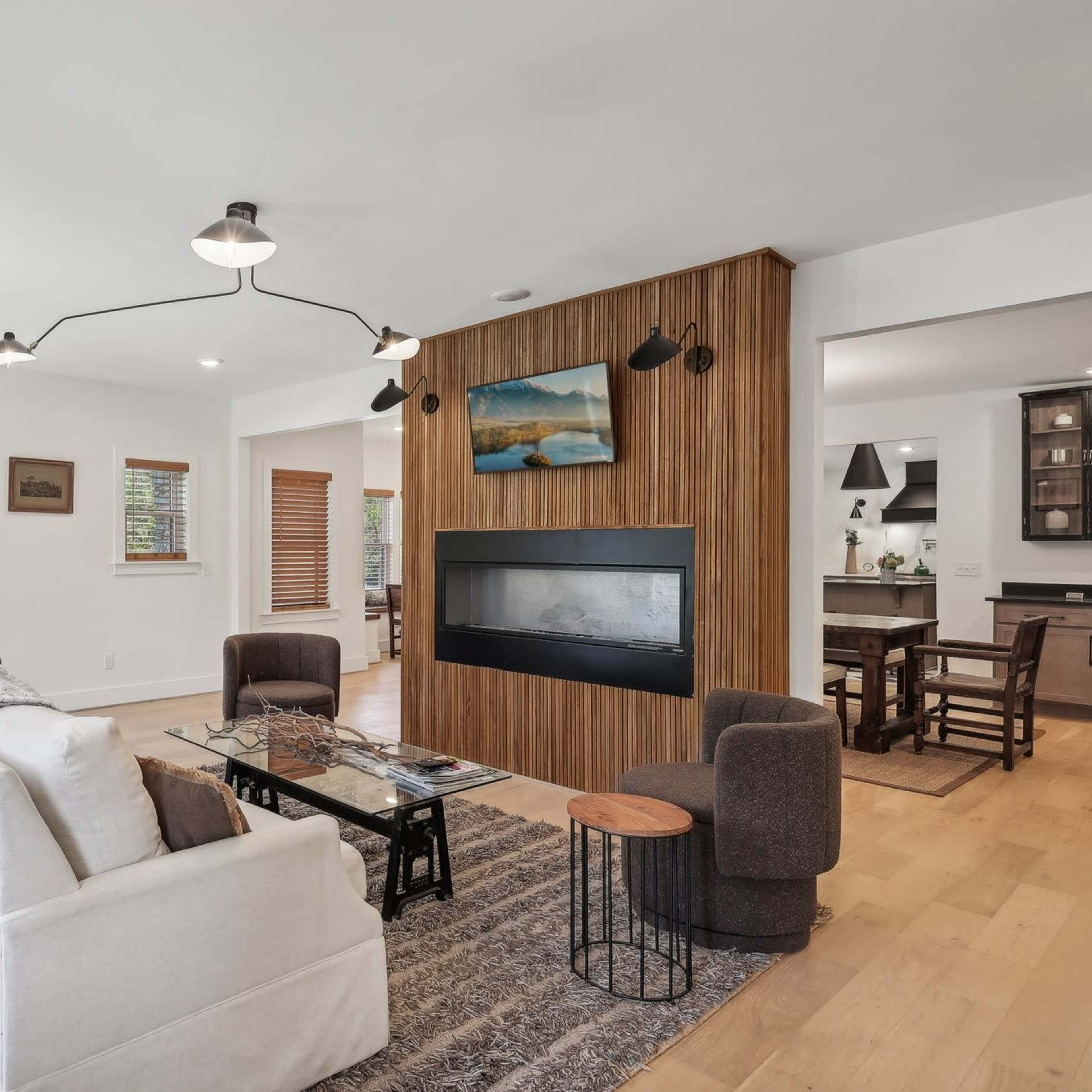 The living room features a modern design with a white sofa, a circular metal side table, and a wall-mounted television above a fireplace, set against wooden paneling.