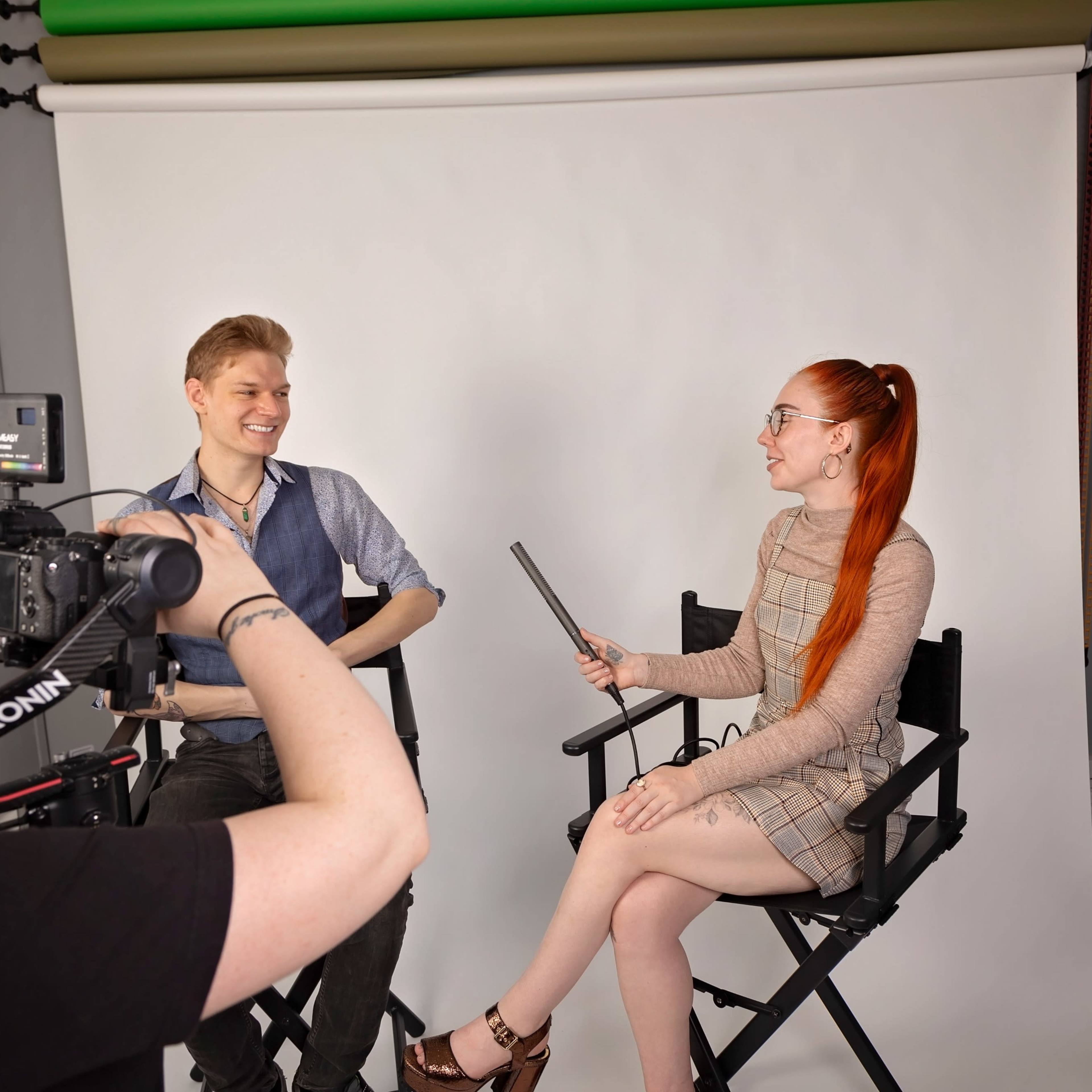 A woman interviews a man in a photography studio, with a camera on a tripod capturing the scene.