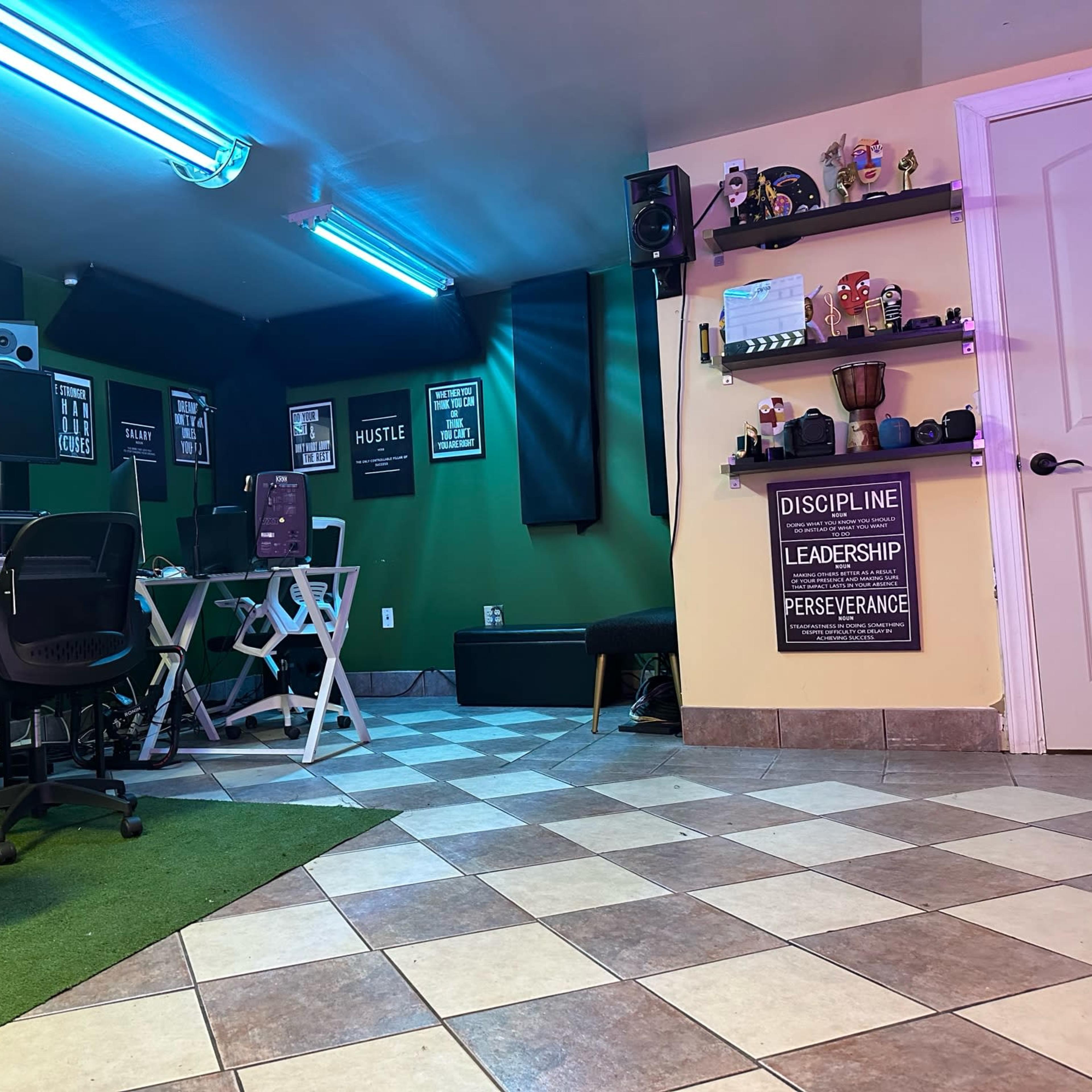 A home office setup with dual computer monitors, speakers, and motivational posters on a green-painted wall, alongside a tiled floor with a small area rug.