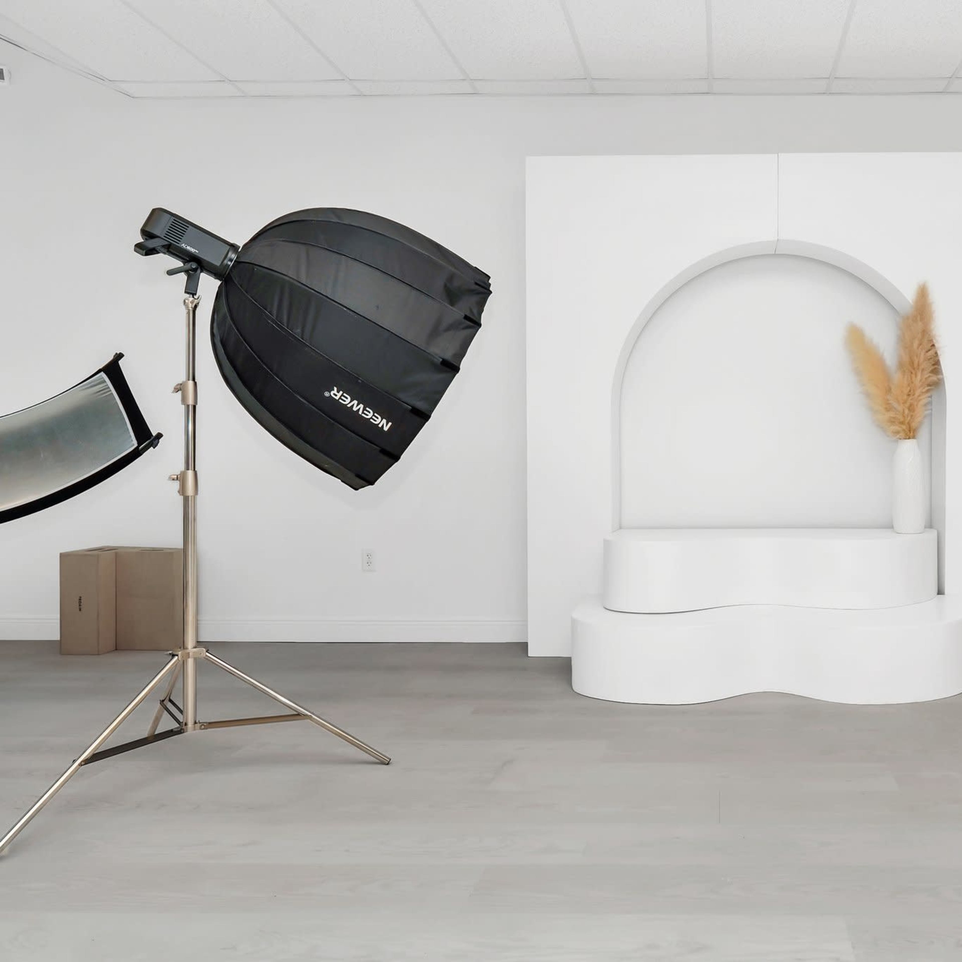 A minimalist studio setup featuring a large black softbox light, a curved white display stand with a plant, and a wooden chair against a plain white wall.