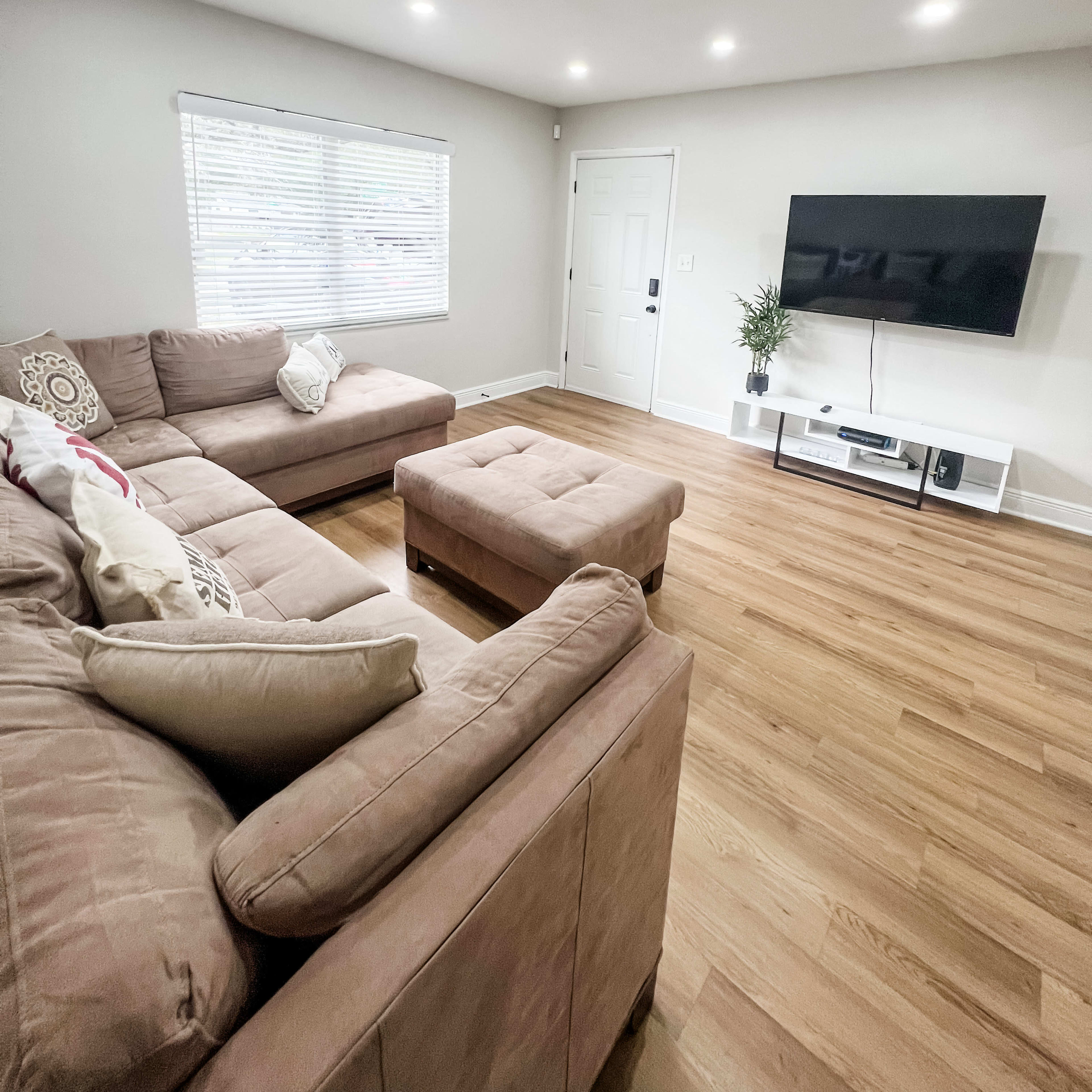 A living room with a large sectional sofa, a coffee table, a television mounted on the wall, and a window with blinds.
