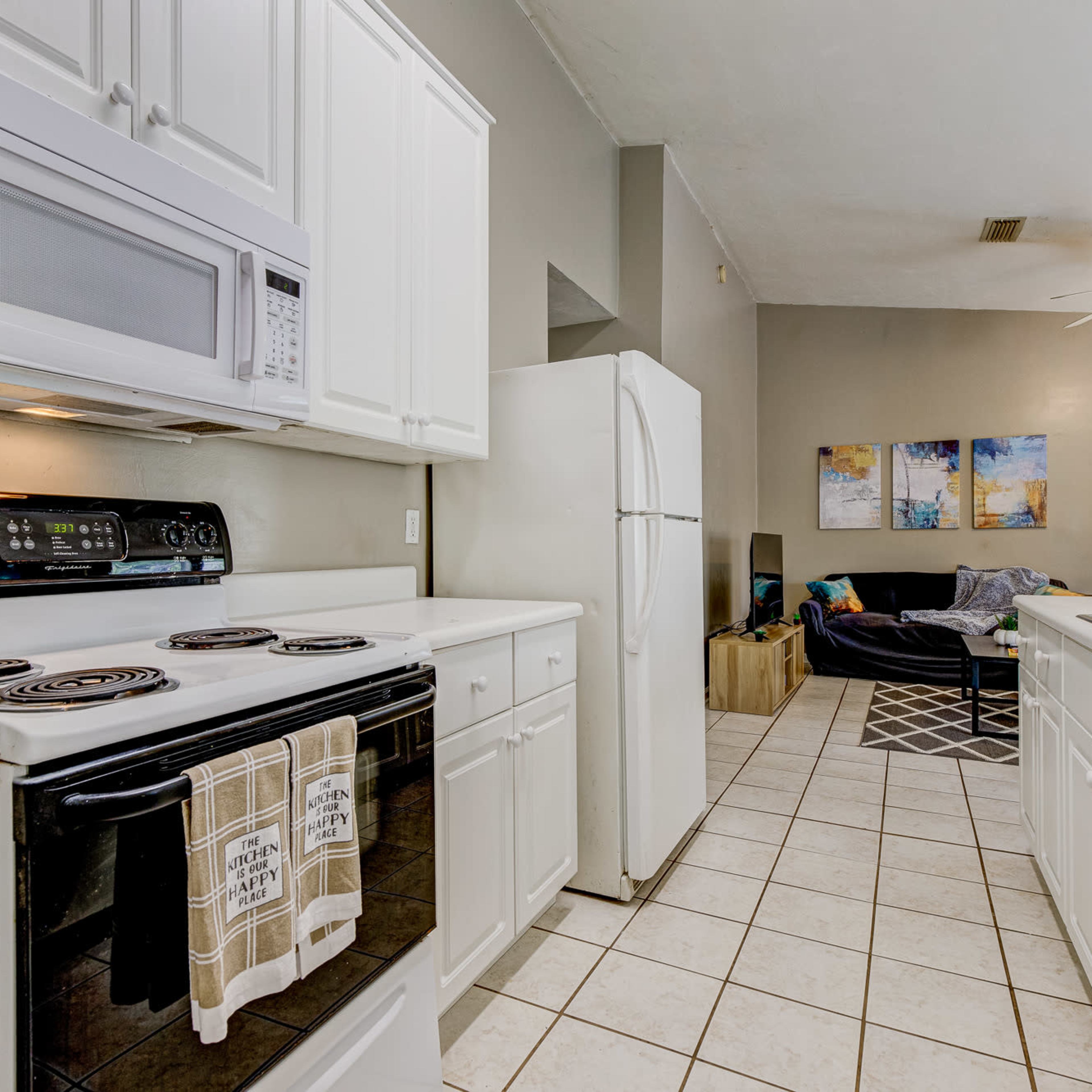 A clean kitchen with white cabinets, a black stove, and a microwave, leading into a living area with a couch and a coffee table.