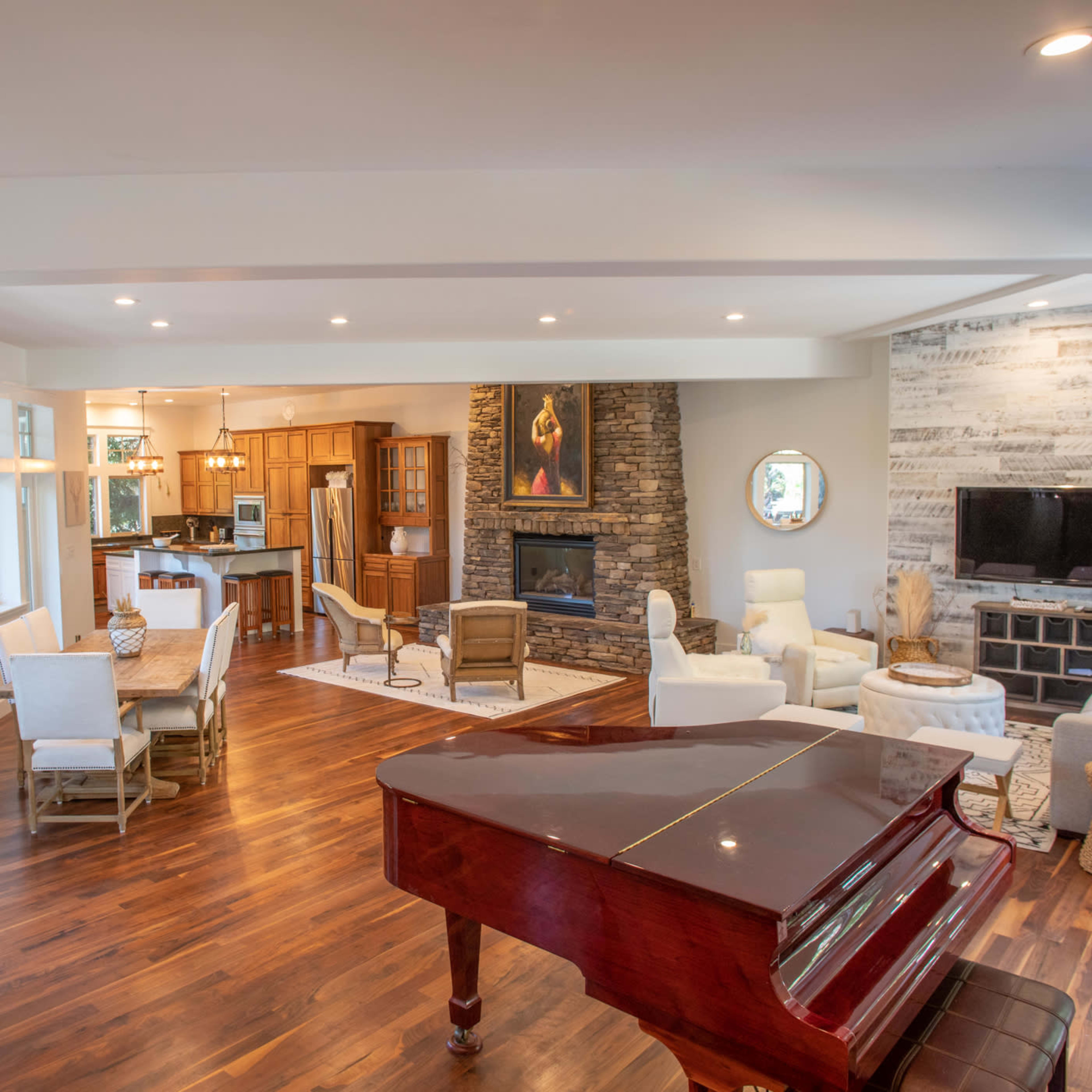 A spacious living room features a grand piano, large windows, a stone fireplace, and an open layout connecting the dining area and kitchen.