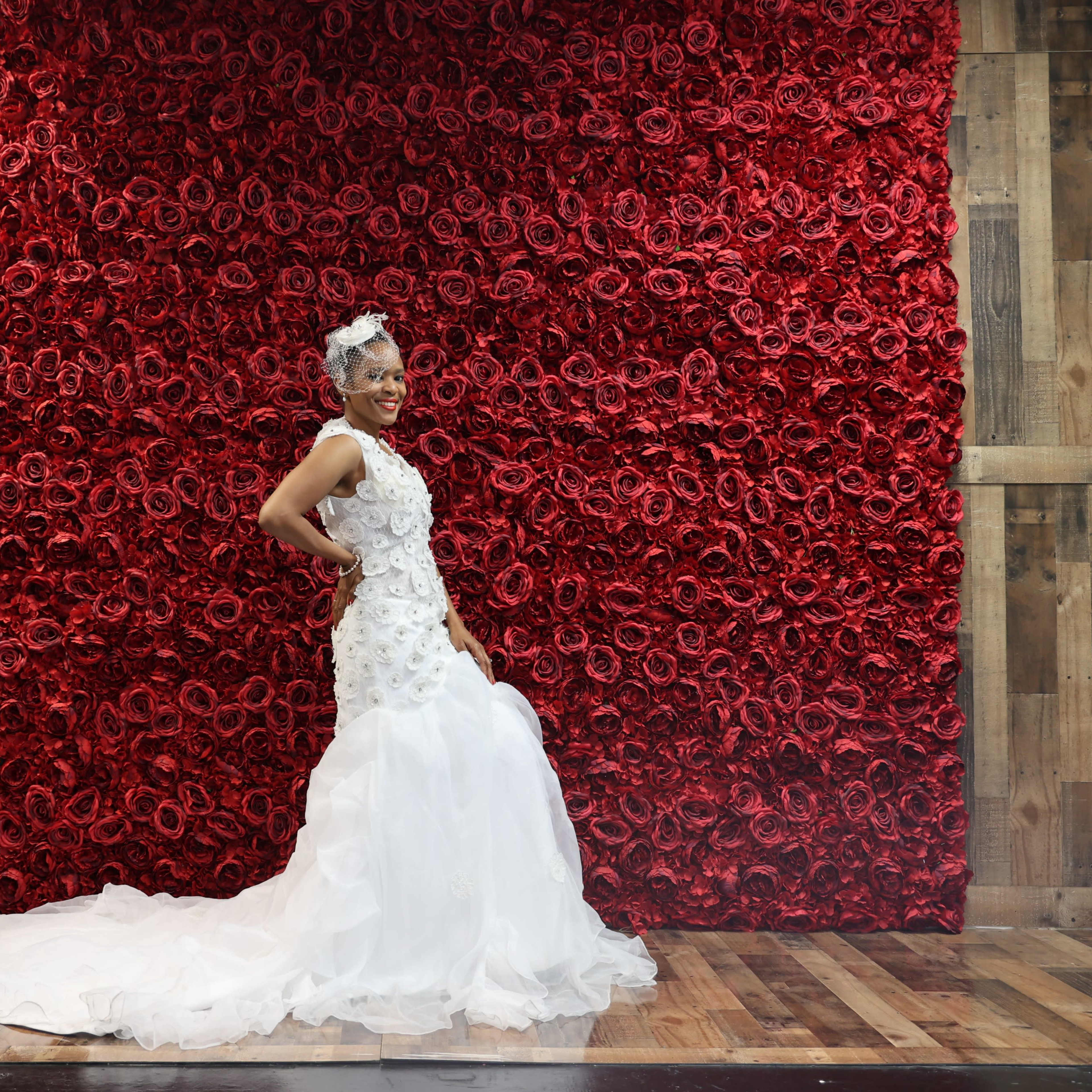 A woman in a white wedding dress poses next to a wall covered in red flowers.