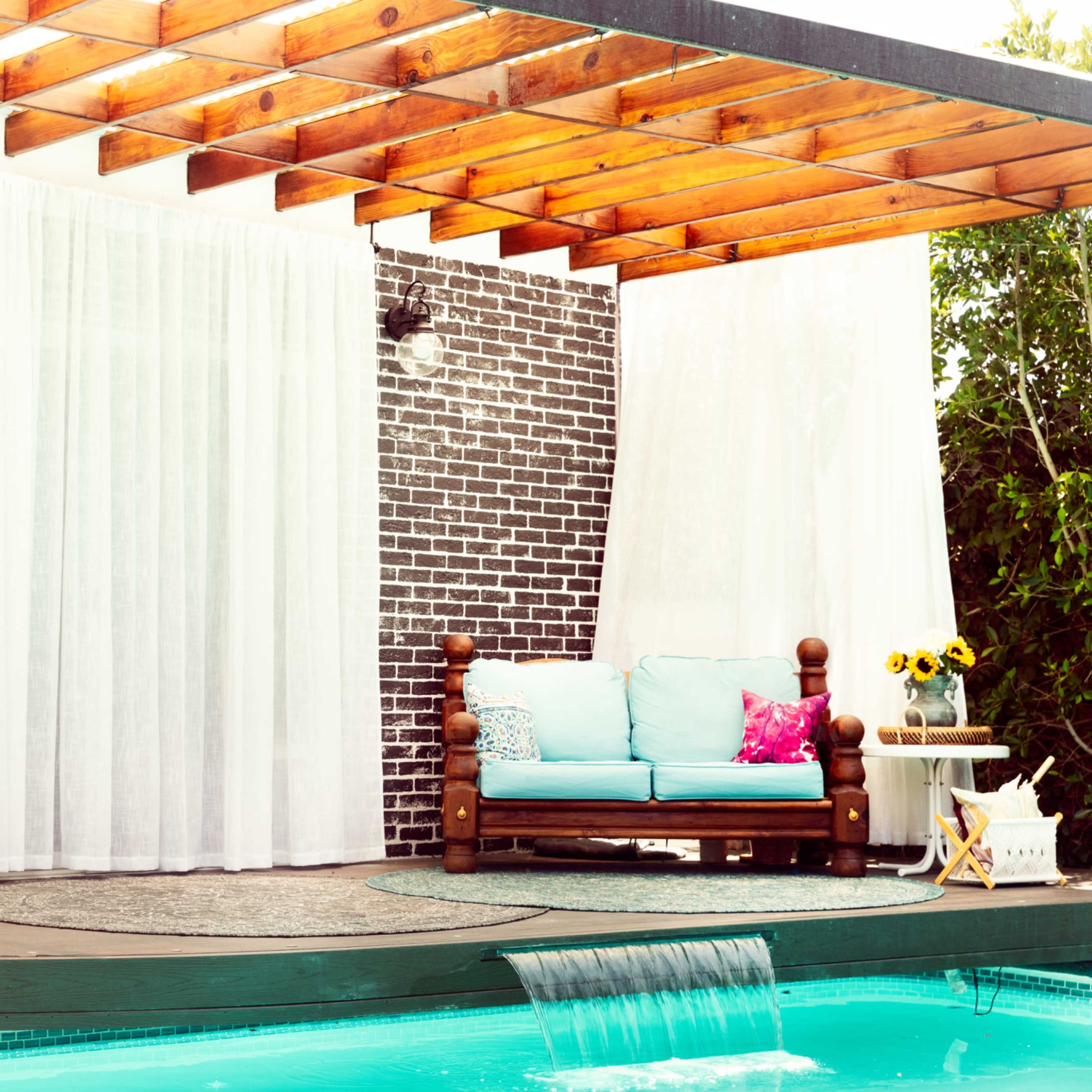 A covered wooden patio with a sofa and decorative pillows overlooks a pool surrounded by a brick wall and greenery.