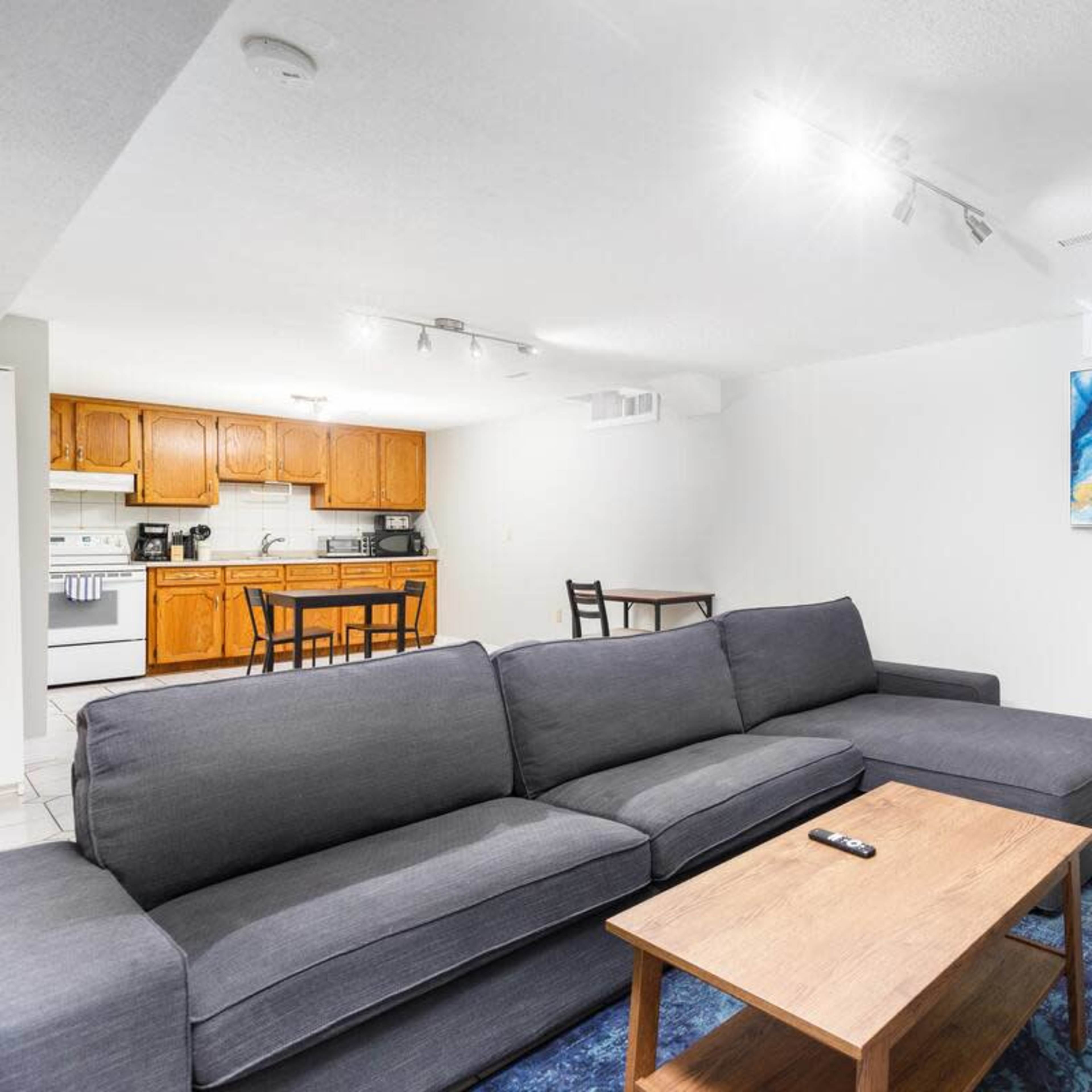 A modern living area with a grey sectional sofa, a wooden coffee table, and a kitchen in the background featuring wooden cabinets and a dining table.