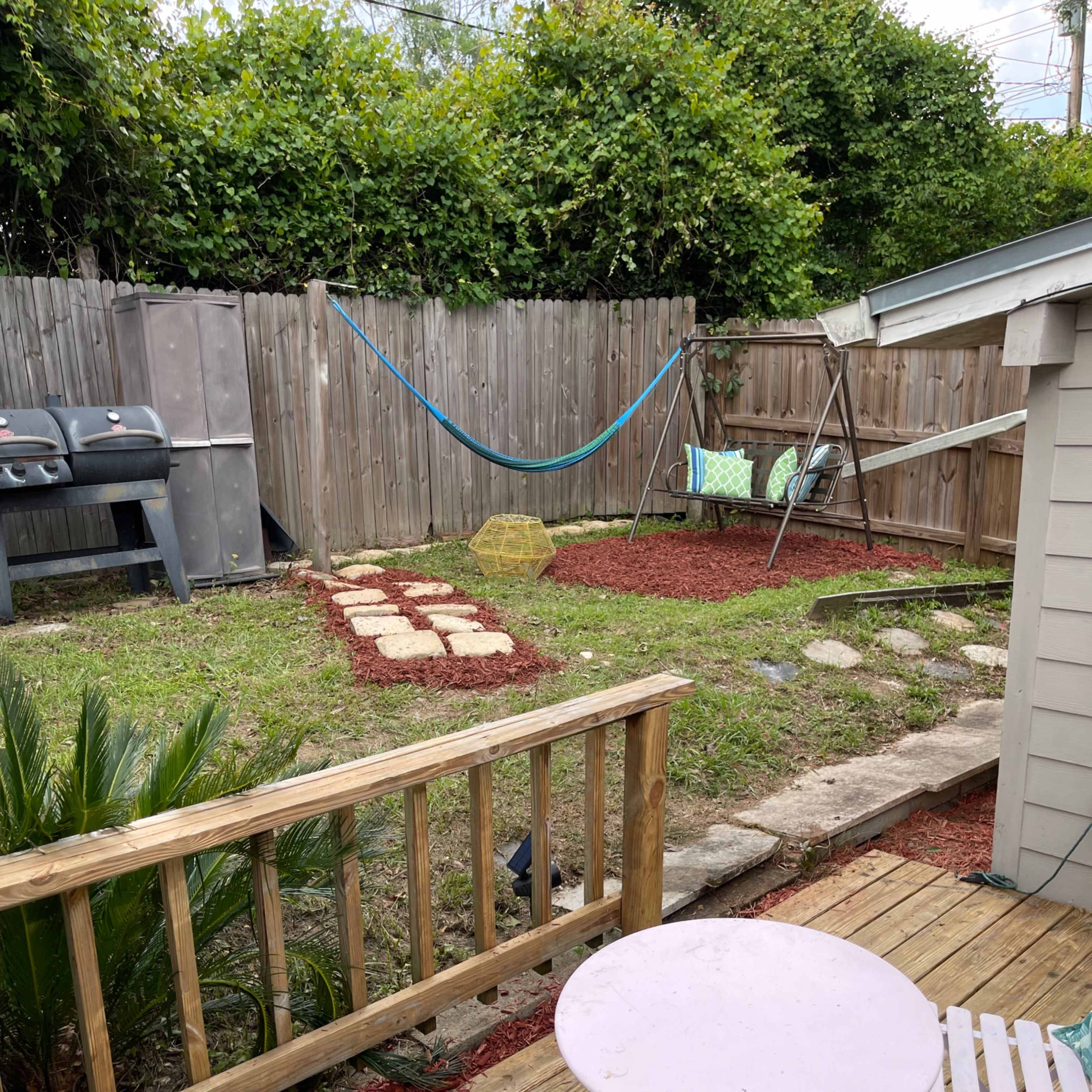 A backyard with a wooden deck, a small table and chairs, a grass area with stepping stones, a hammock, a swing set, and a grill against a wooden fence.