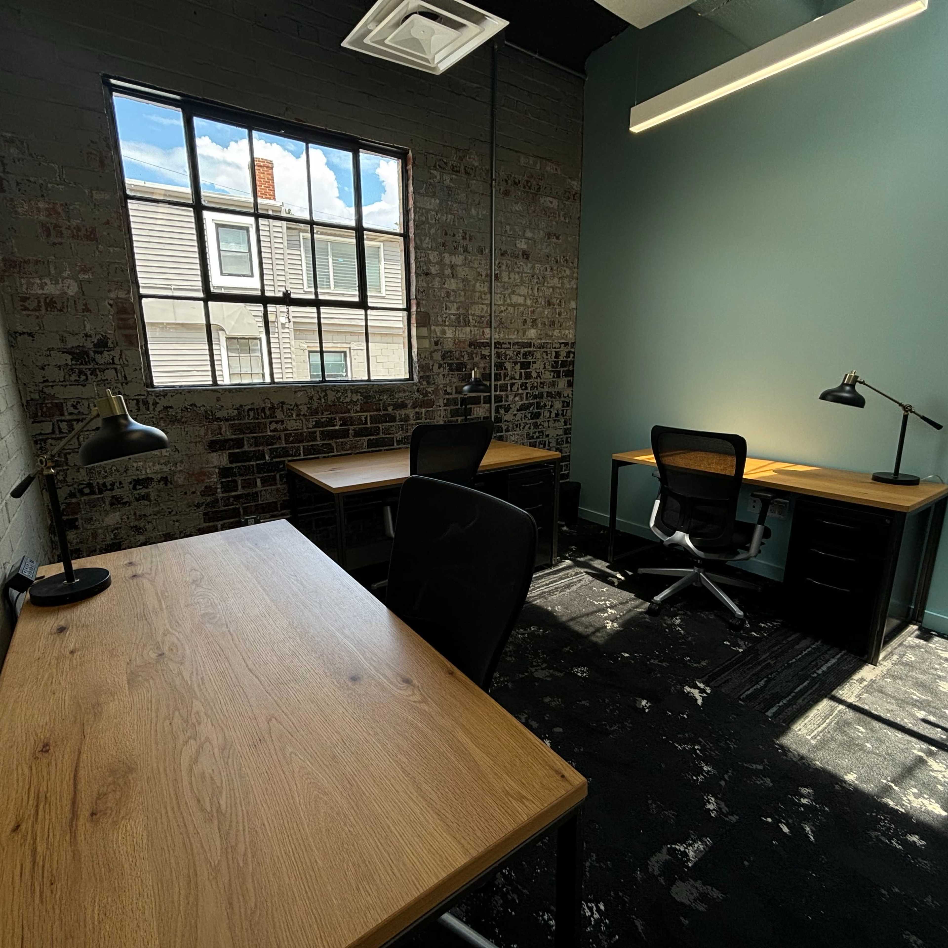 A well-lit office space with two wooden desks and a chair, featuring exposed brick walls and a large window that opens to an exterior view.