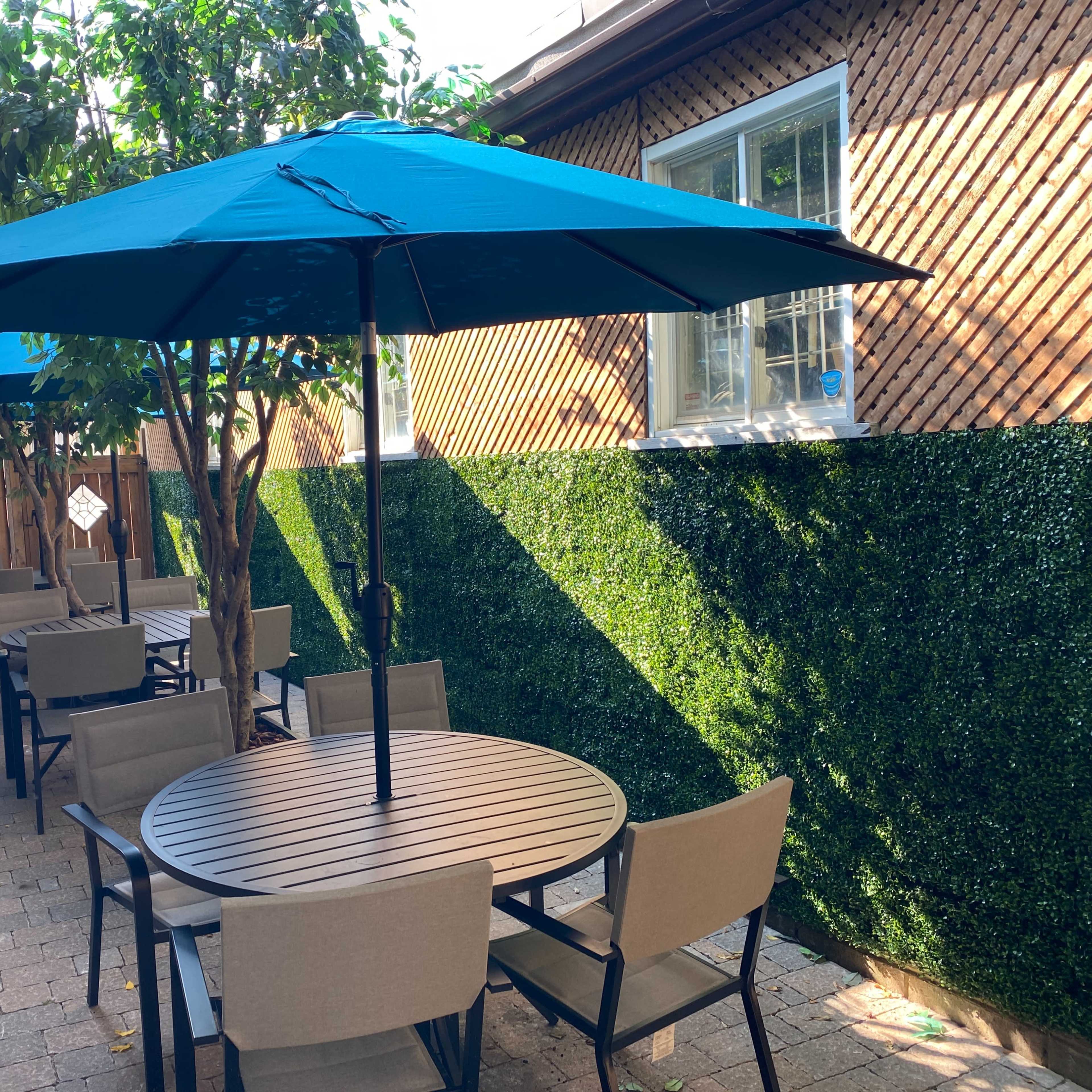 A courtyard features several round tables with beige chairs beneath large blue umbrellas, surrounded by green hedges and brick walls.
