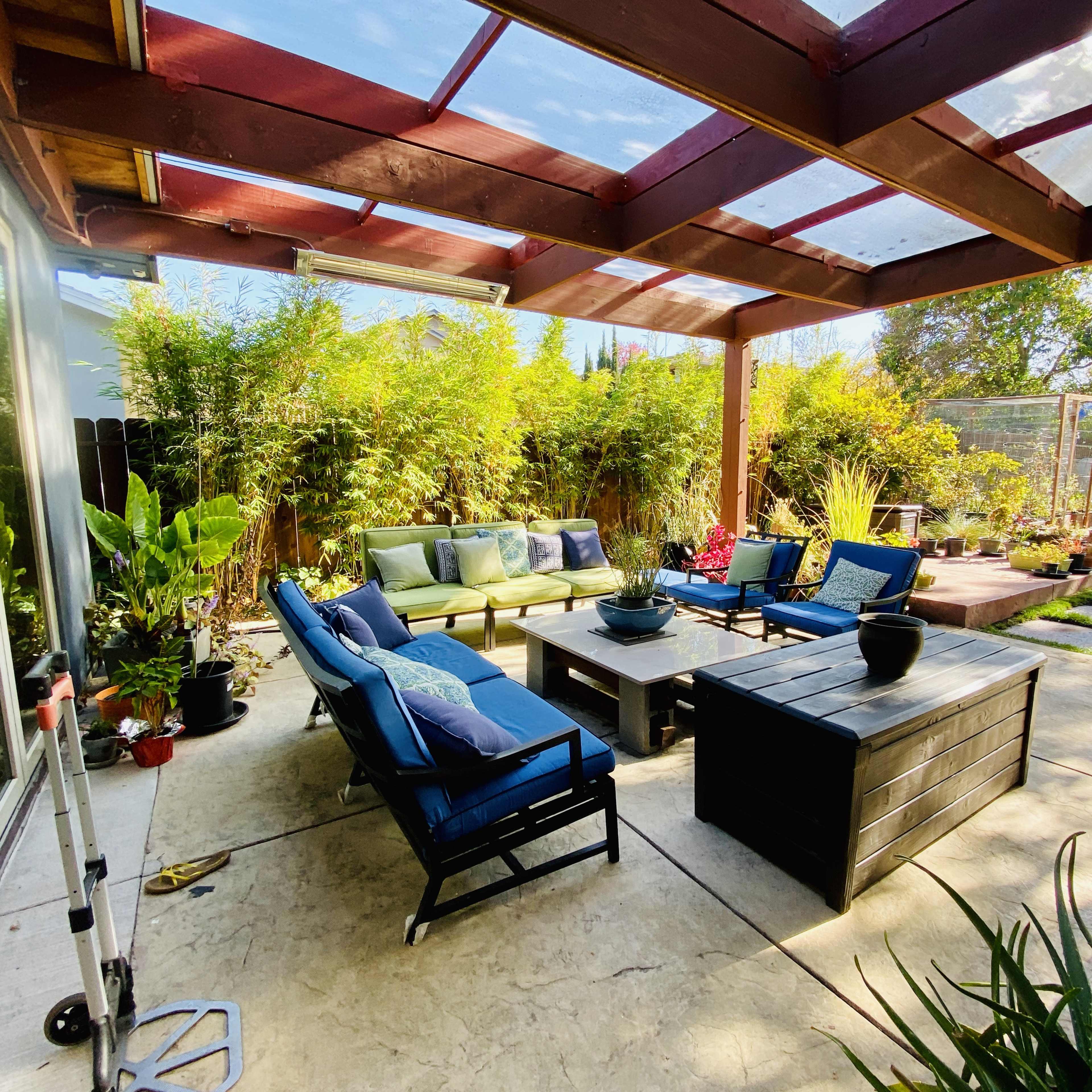 A covered patio features a seating area with blue and green cushions surrounding a central table, complemented by various plants and a stone pathway in the background.