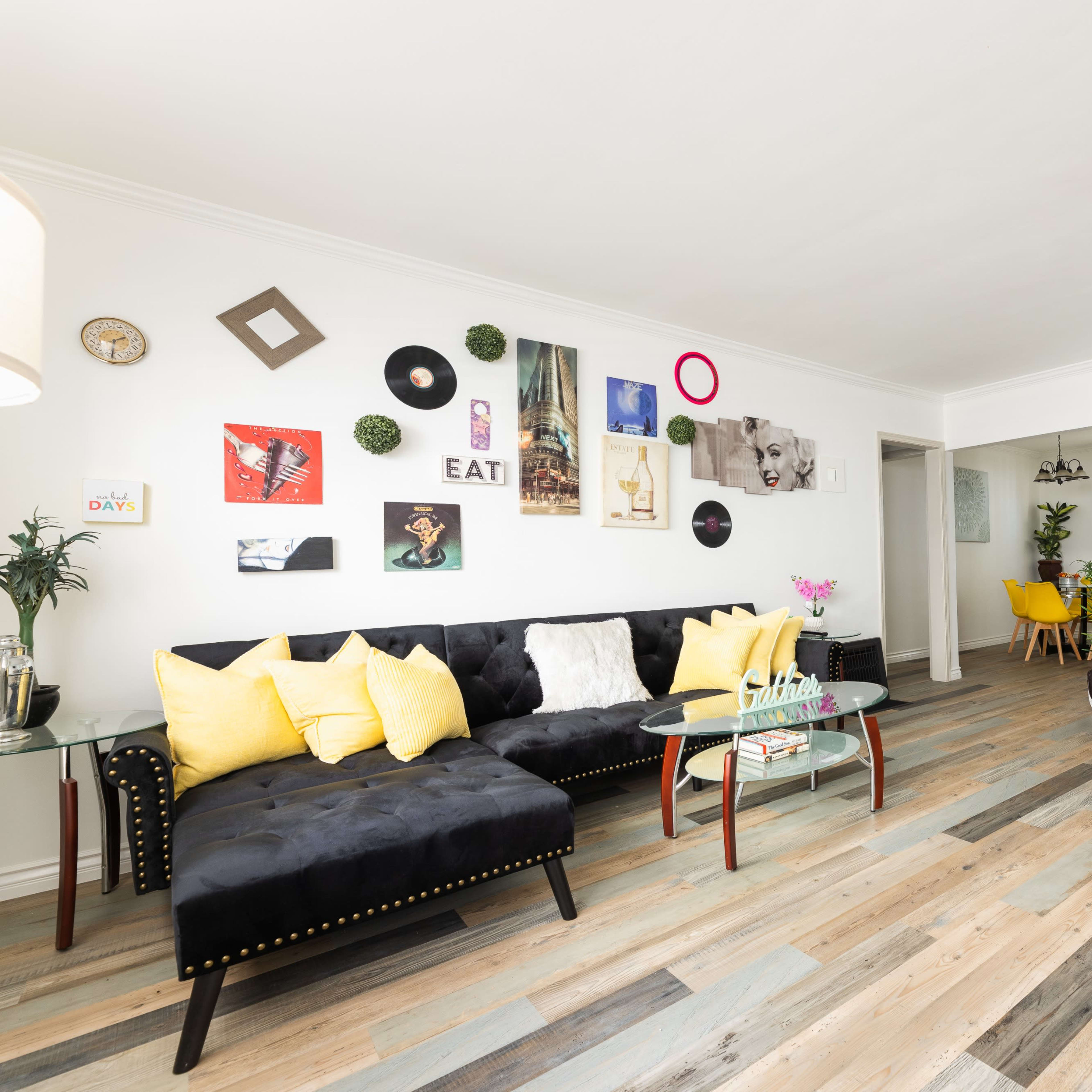 The living room features two sofas, a glass coffee table, and a decorative wall adorned with various artwork and plants.