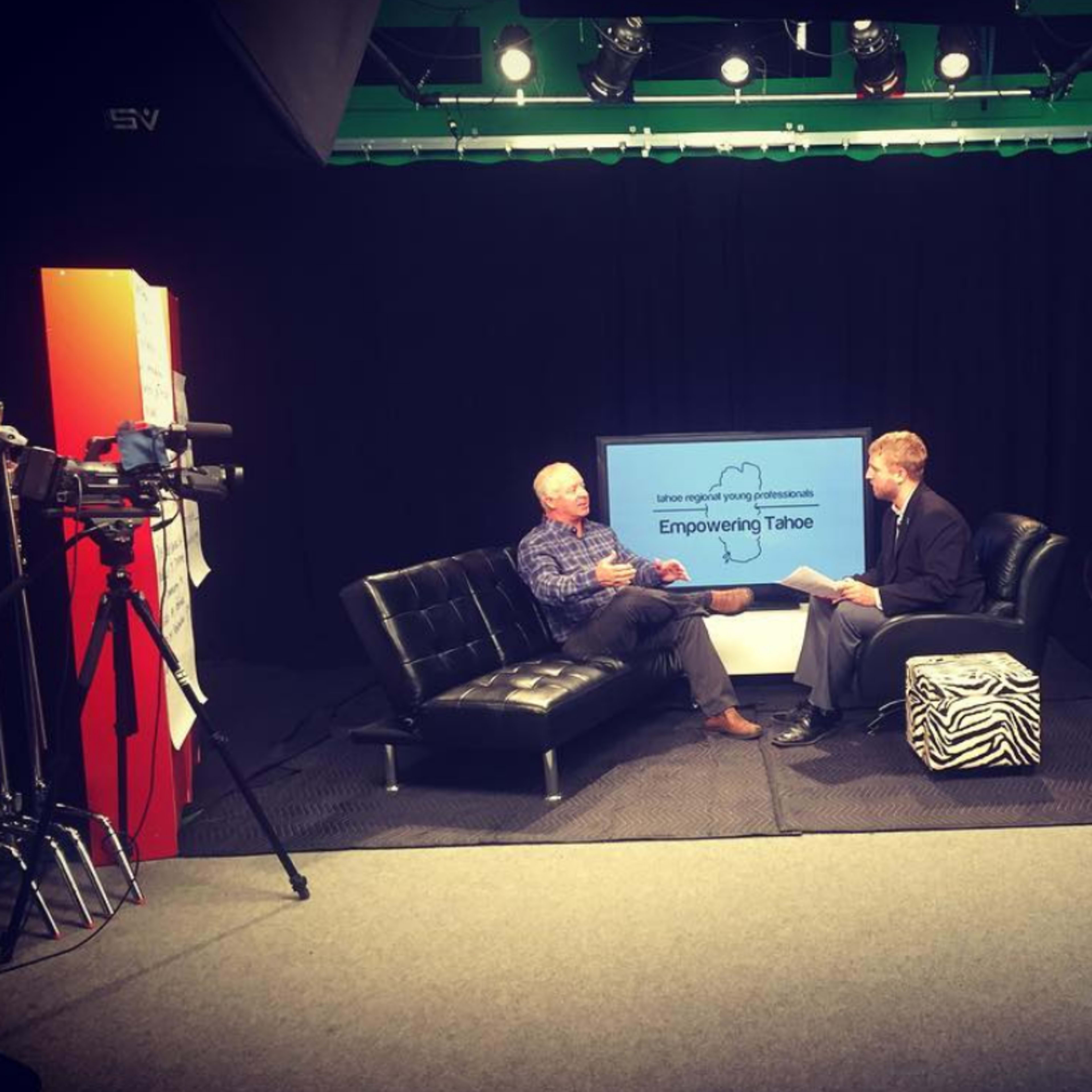 A television studio features two men seated on a couch discussing "Empowering Tahoe," with cameras and lights positioned around them.