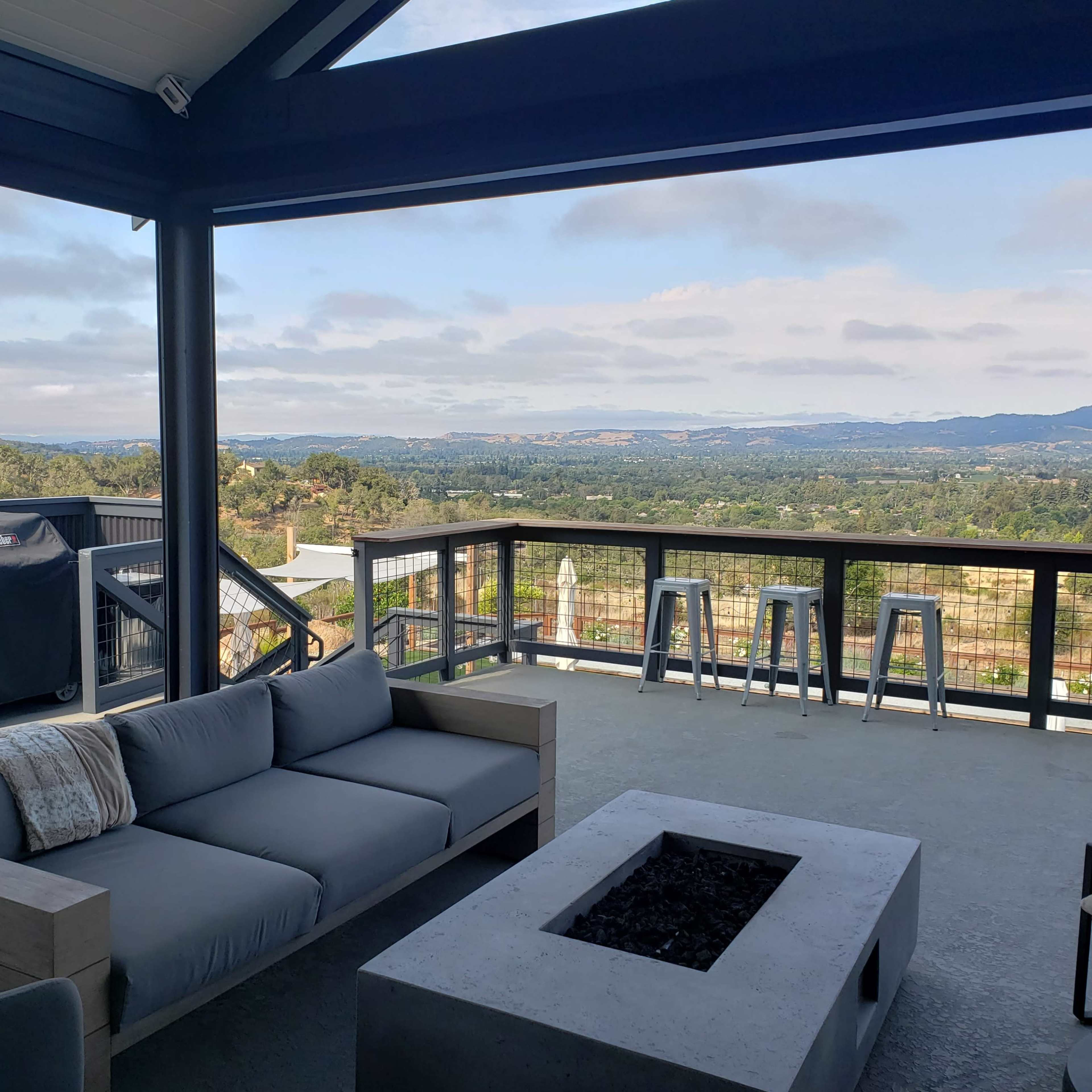 A modern outdoor patio features a gray sofa and a fire pit with a view of rolling hills and distant mountains under a cloudy sky.