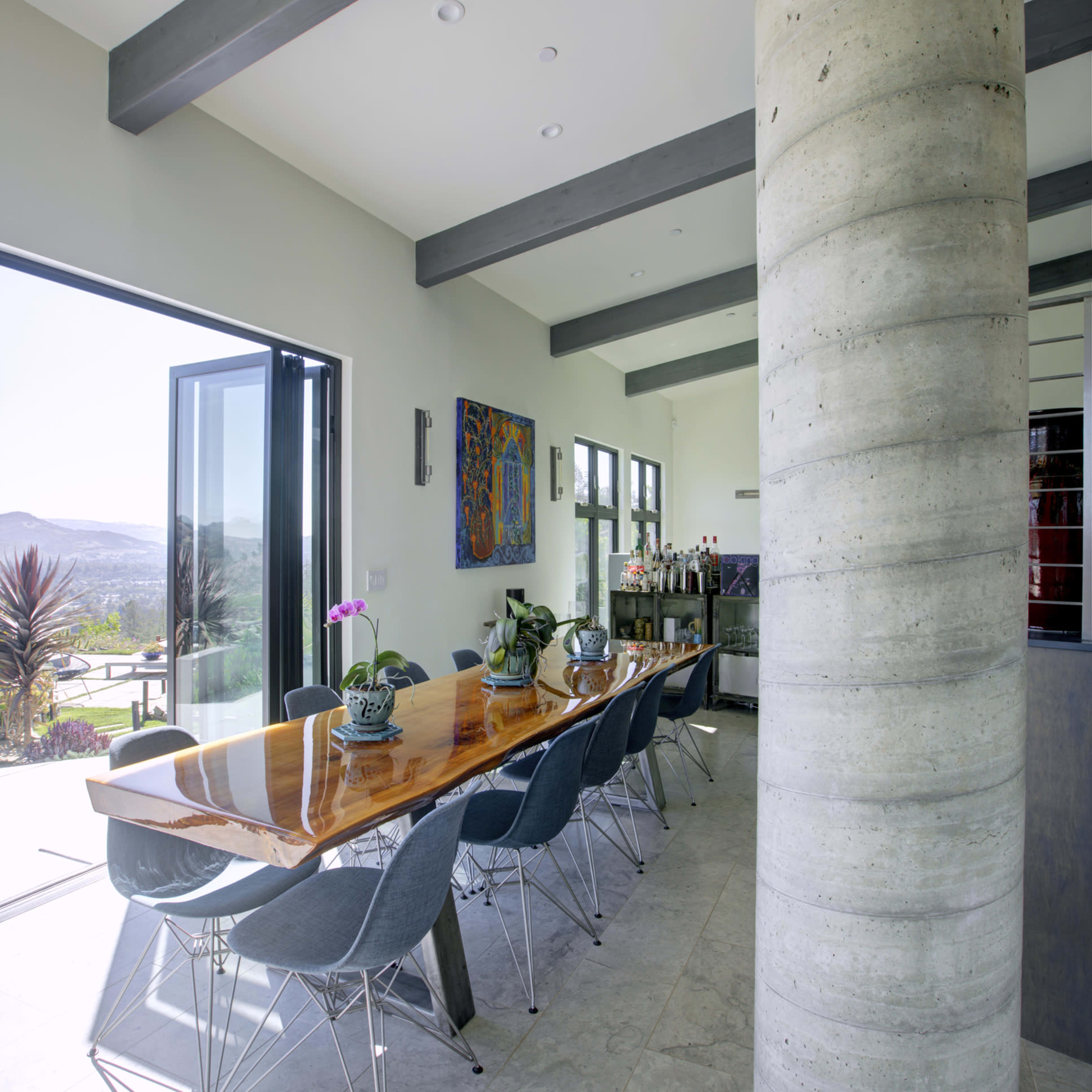 A long wooden dining table with modern chairs is set in a brightly lit room with large windows and a concrete column.