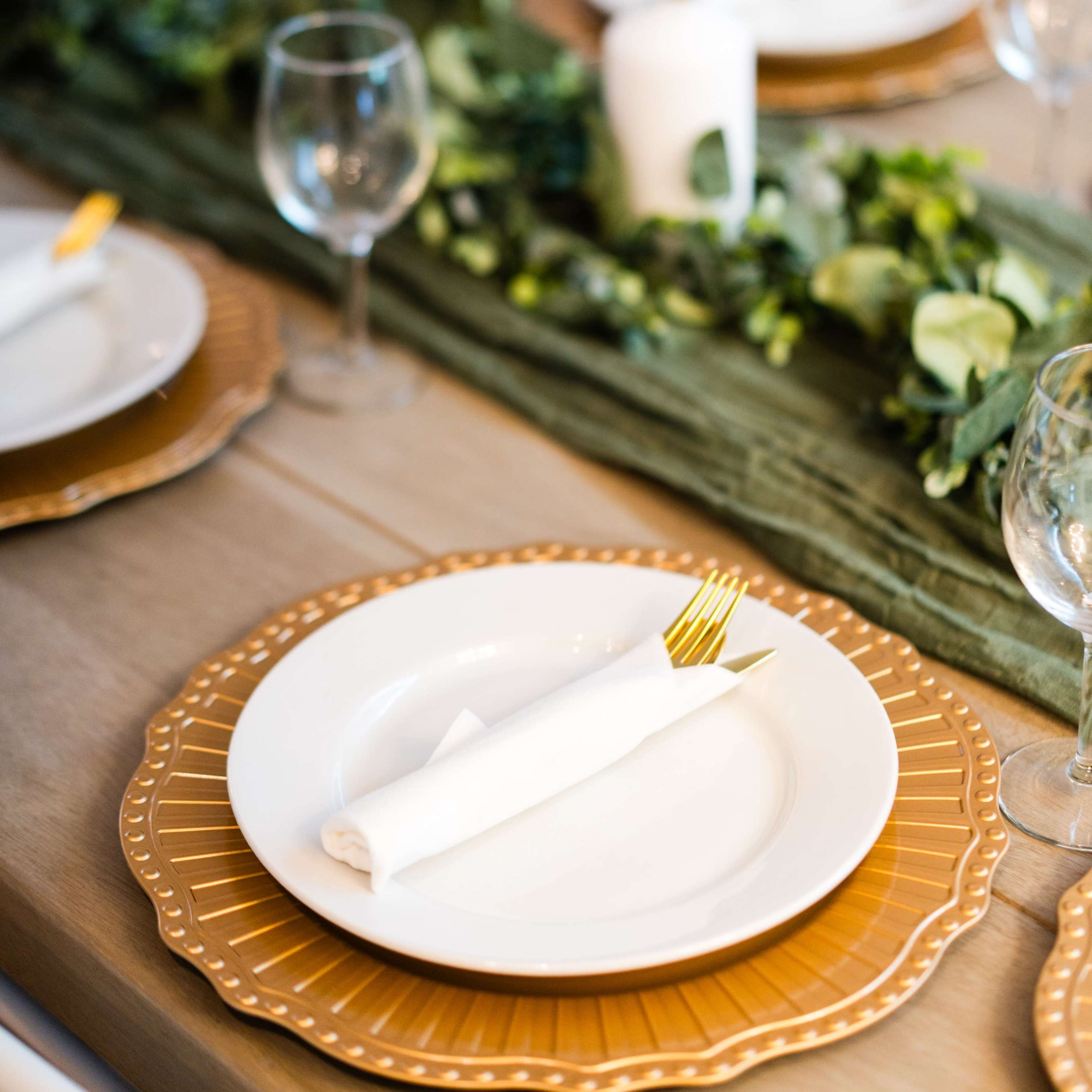 A dining table is elegantly set with white plates, golden chargers, wine glasses, and candles, accented by a green centerpiece.