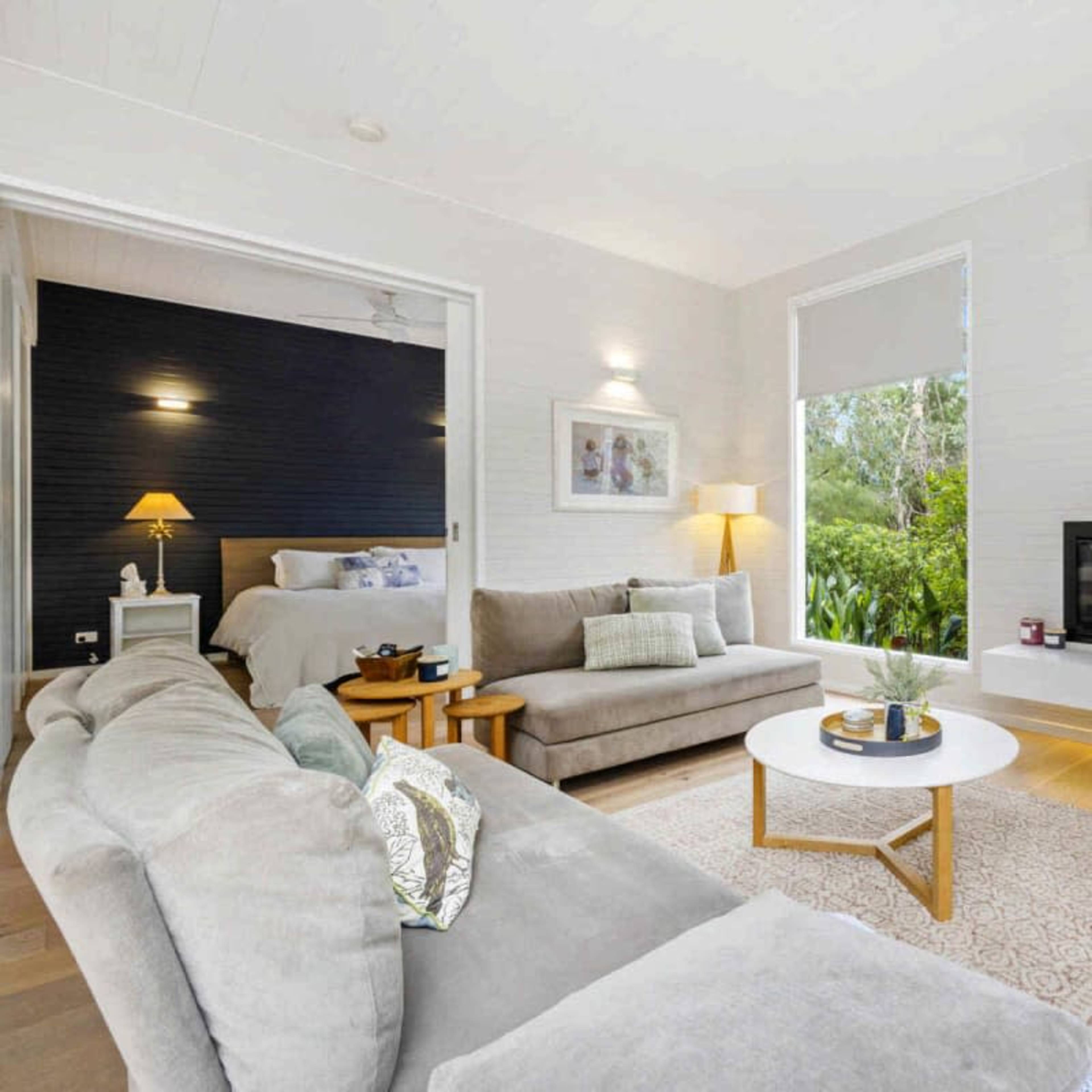 A modern living room with a gray sofa, a circular coffee table, a flat-screen TV, and a view of a bedroom area through an open doorway, featuring a bed with a lamp.