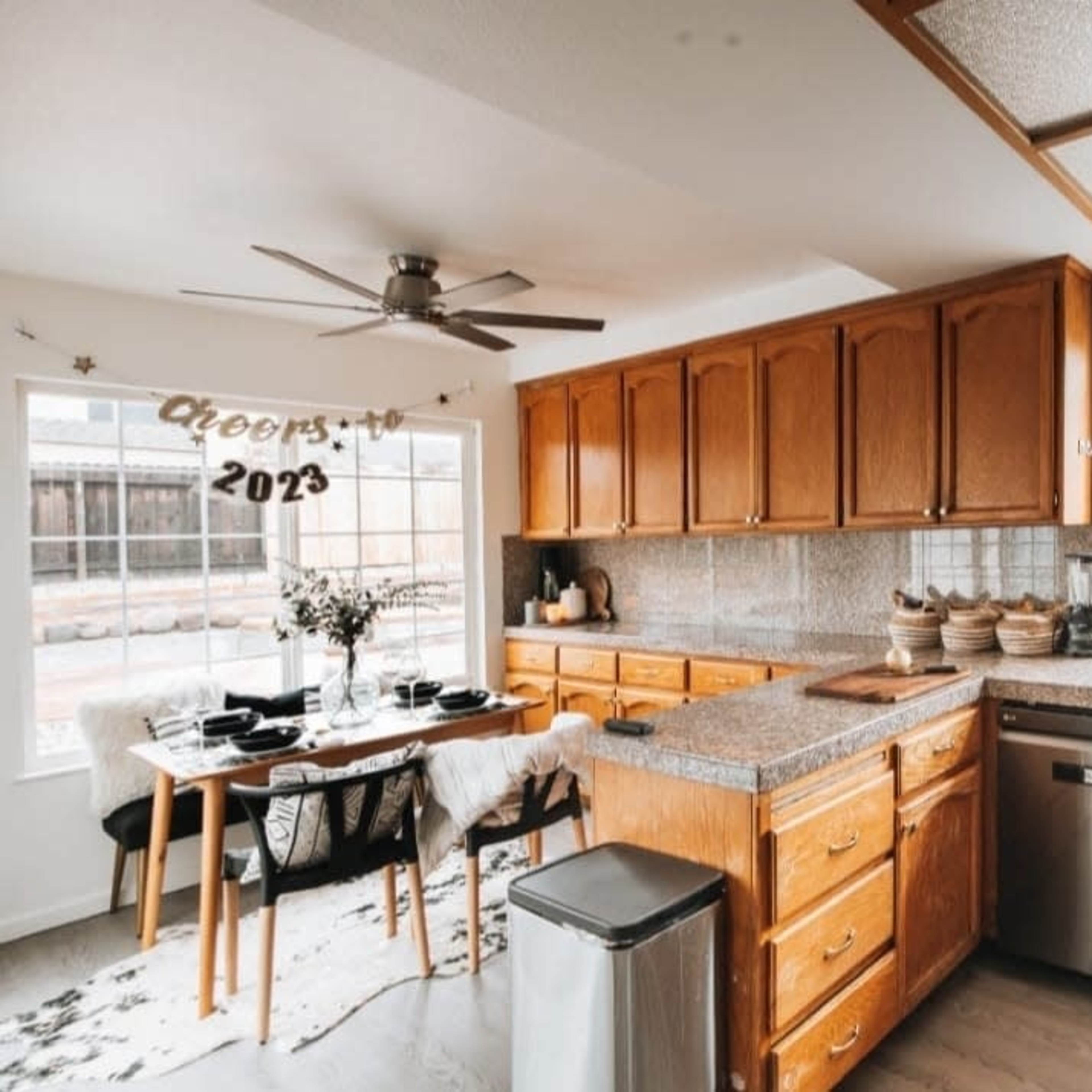 A modern kitchen with wooden cabinets, a granite countertop, a dining table set for four, and large windows allowing natural light.