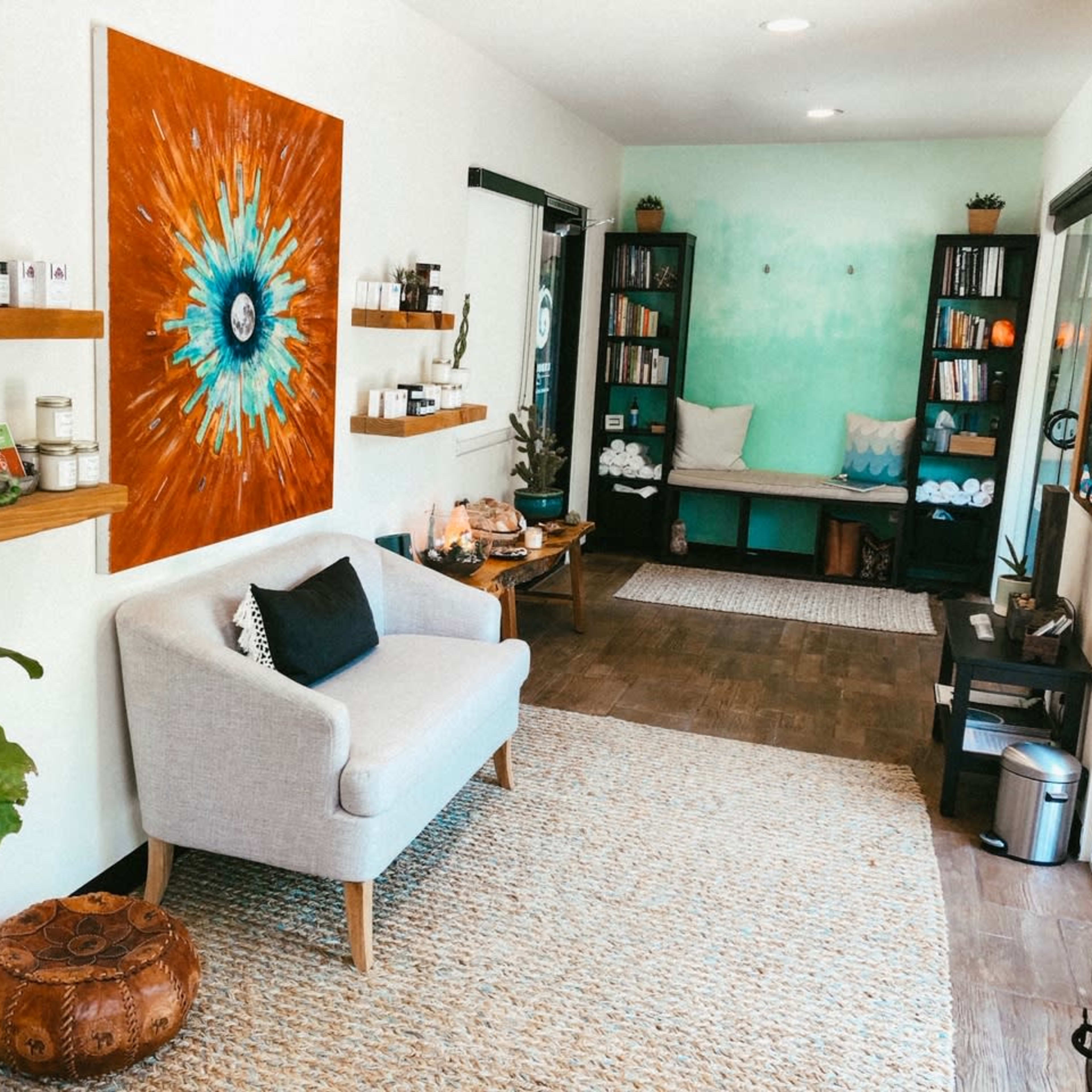 A cozy wellness space featuring a grey armchair, a large vibrant artwork, wooden shelves lined with jars and books, and a seating area with plants and decorative elements.