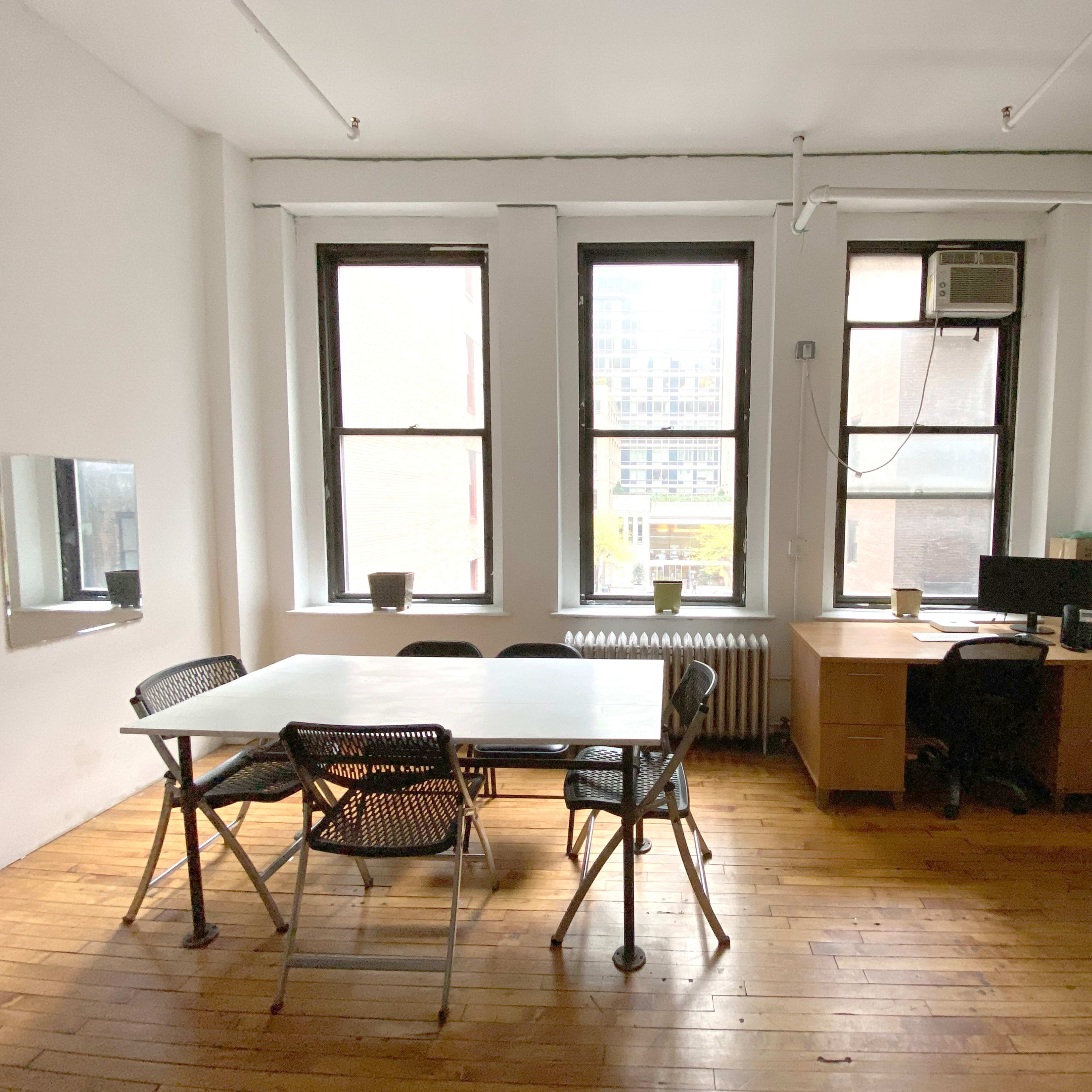 A bright, empty room with wooden floors, a large table surrounded by chairs, a desk with a computer, and windows letting in natural light.