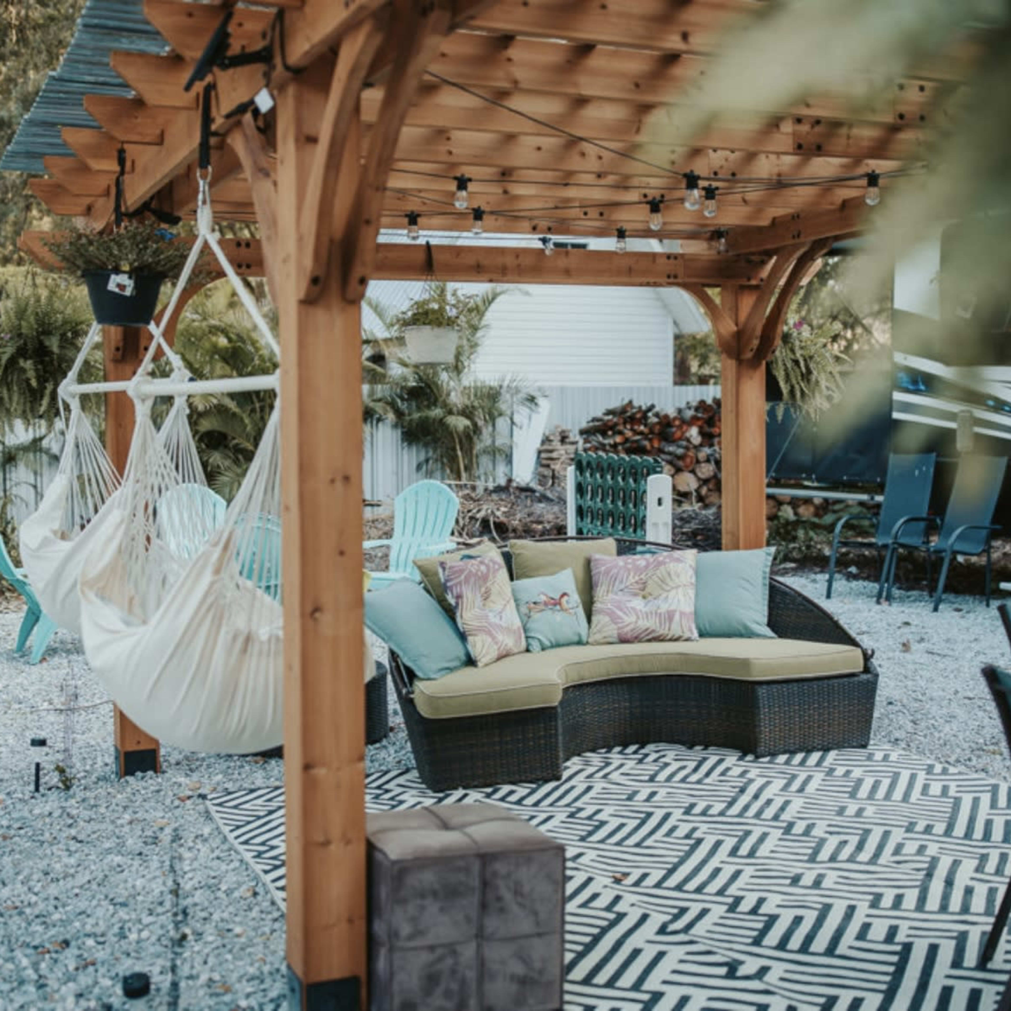 A wooden pergola with a hammock and a sectional sofa is set on a gravel patio with decorative plants and outdoor furniture.
