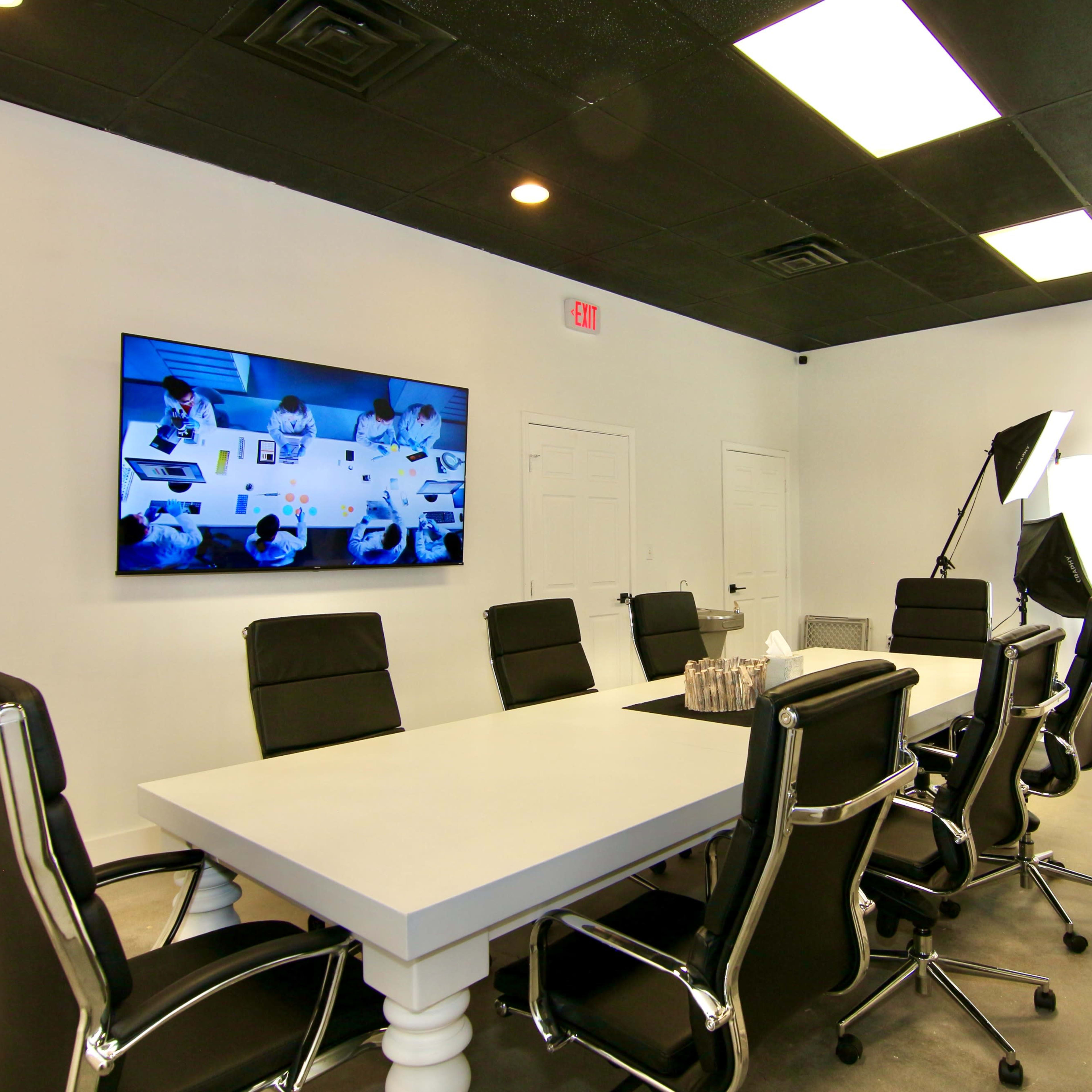 A conference room features a long white table with black chairs, a large screen displaying a meeting, and professional lighting equipment in the corner.