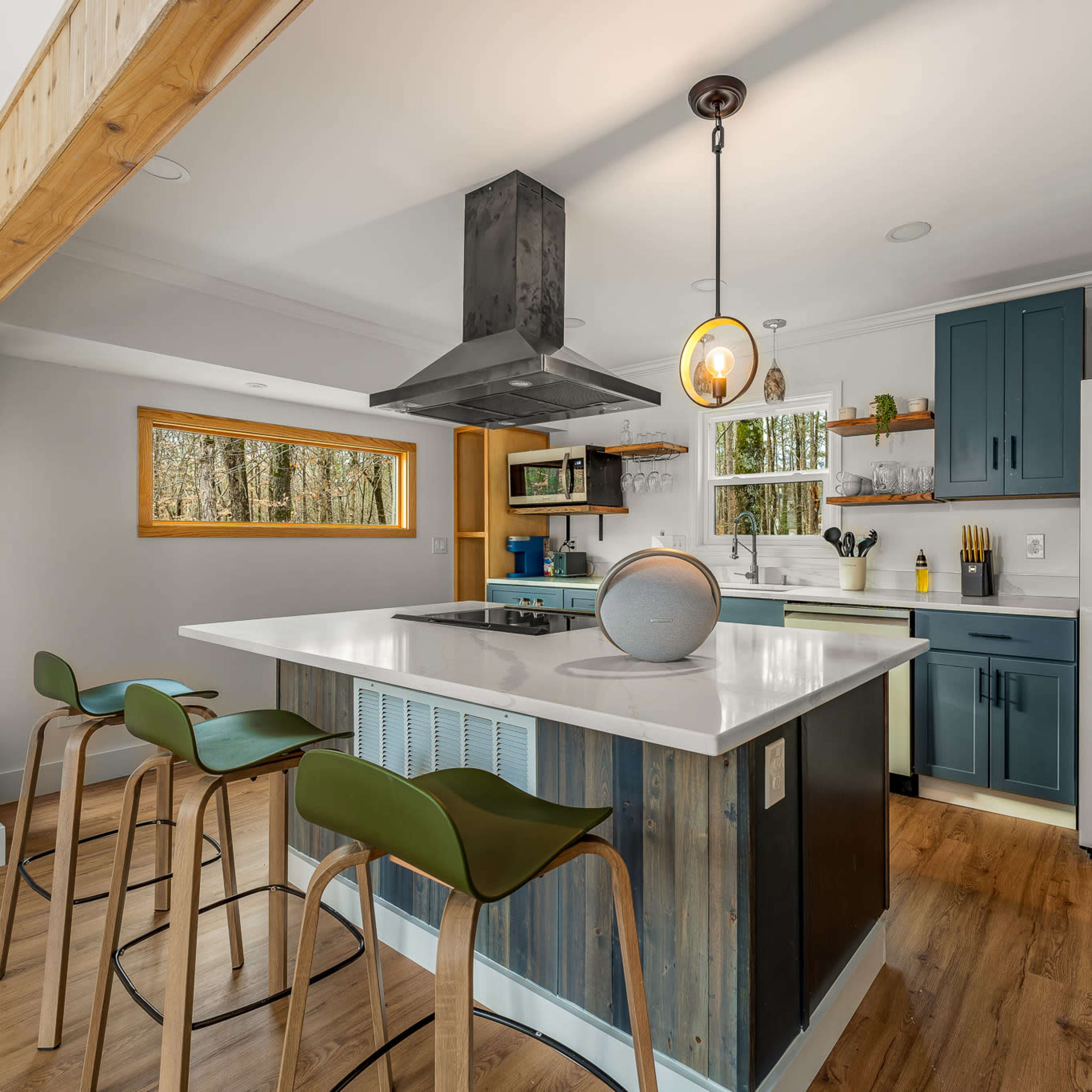 The kitchen features a central island with bar seating, a metal range hood, and blue cabinets against a backdrop of large windows.
