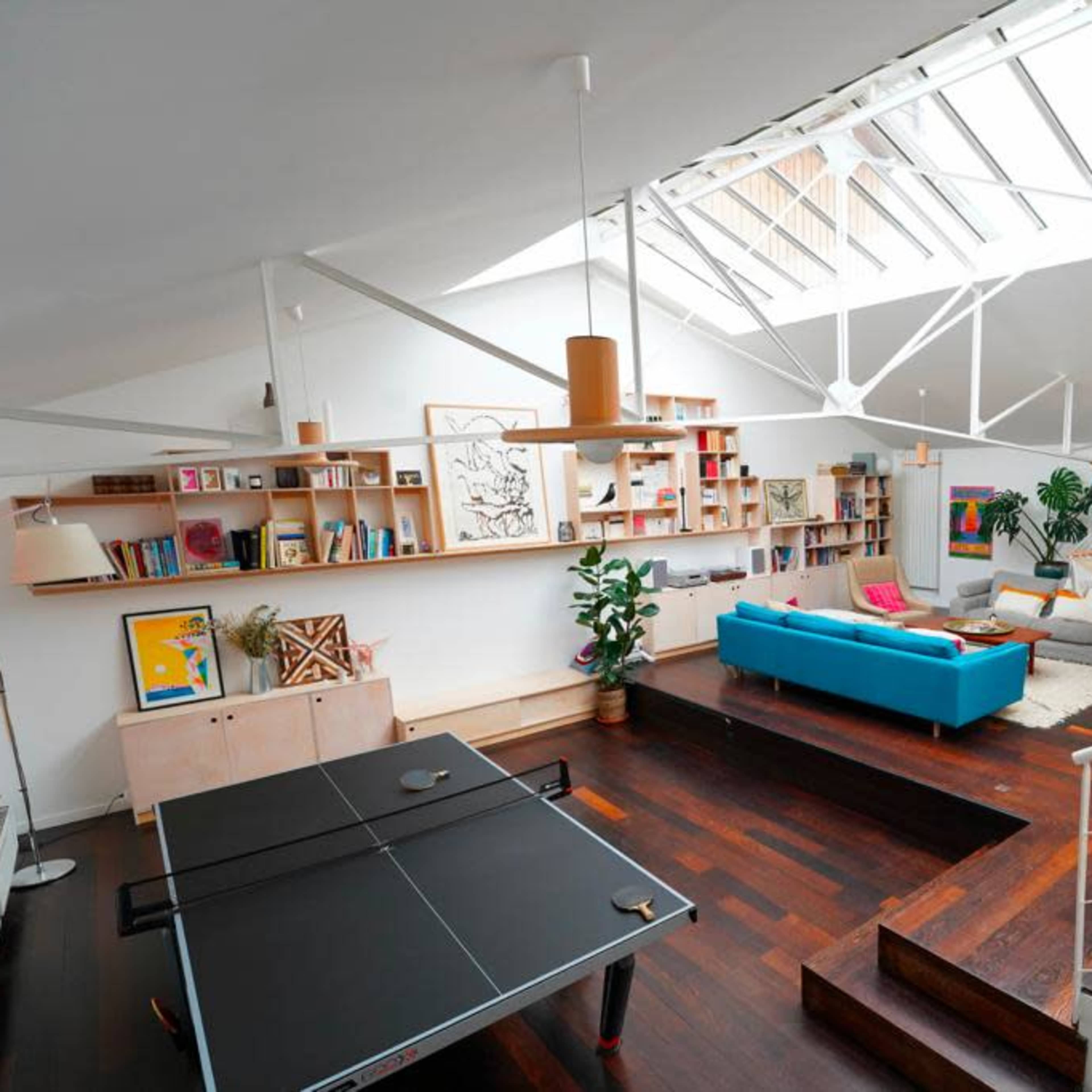 A spacious, modern living area with a skylight, bookshelves, a blue couch, a table tennis setup, and various plants.