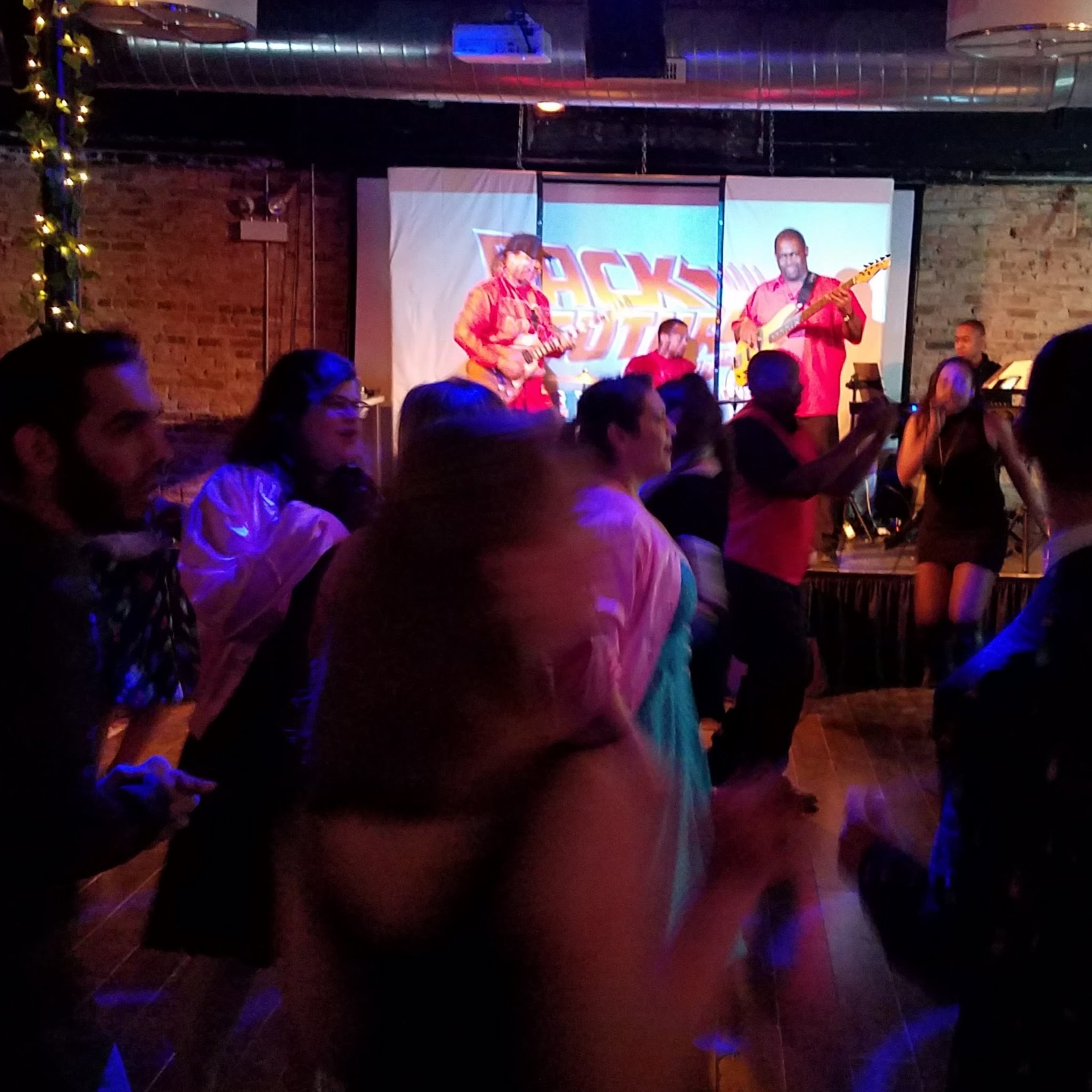 A lively crowd dances in front of a band performing on a stage with colorful lighting.