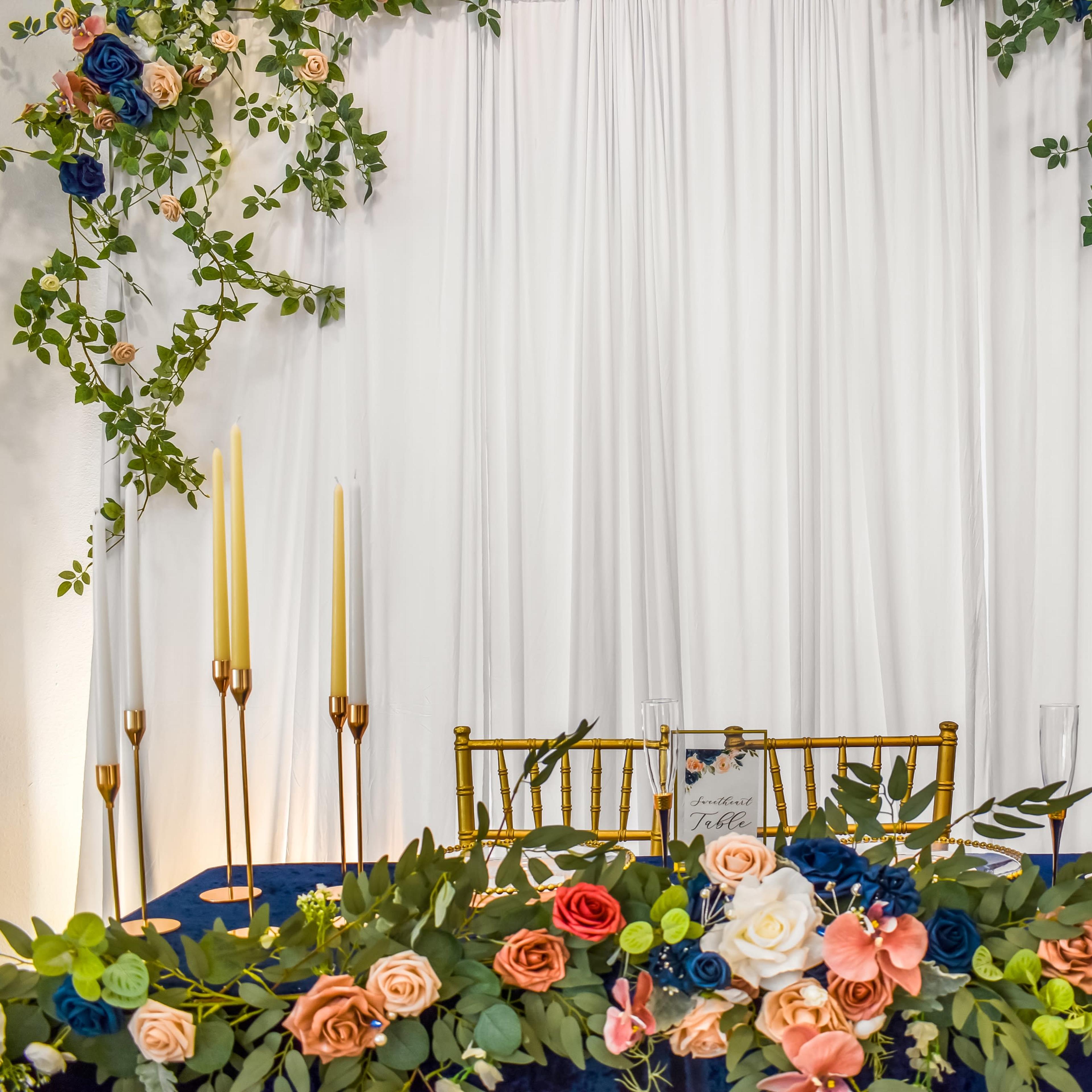 A decorated table with a floral arrangement and tall candle holders stands against a backdrop of draped white fabric and greenery.