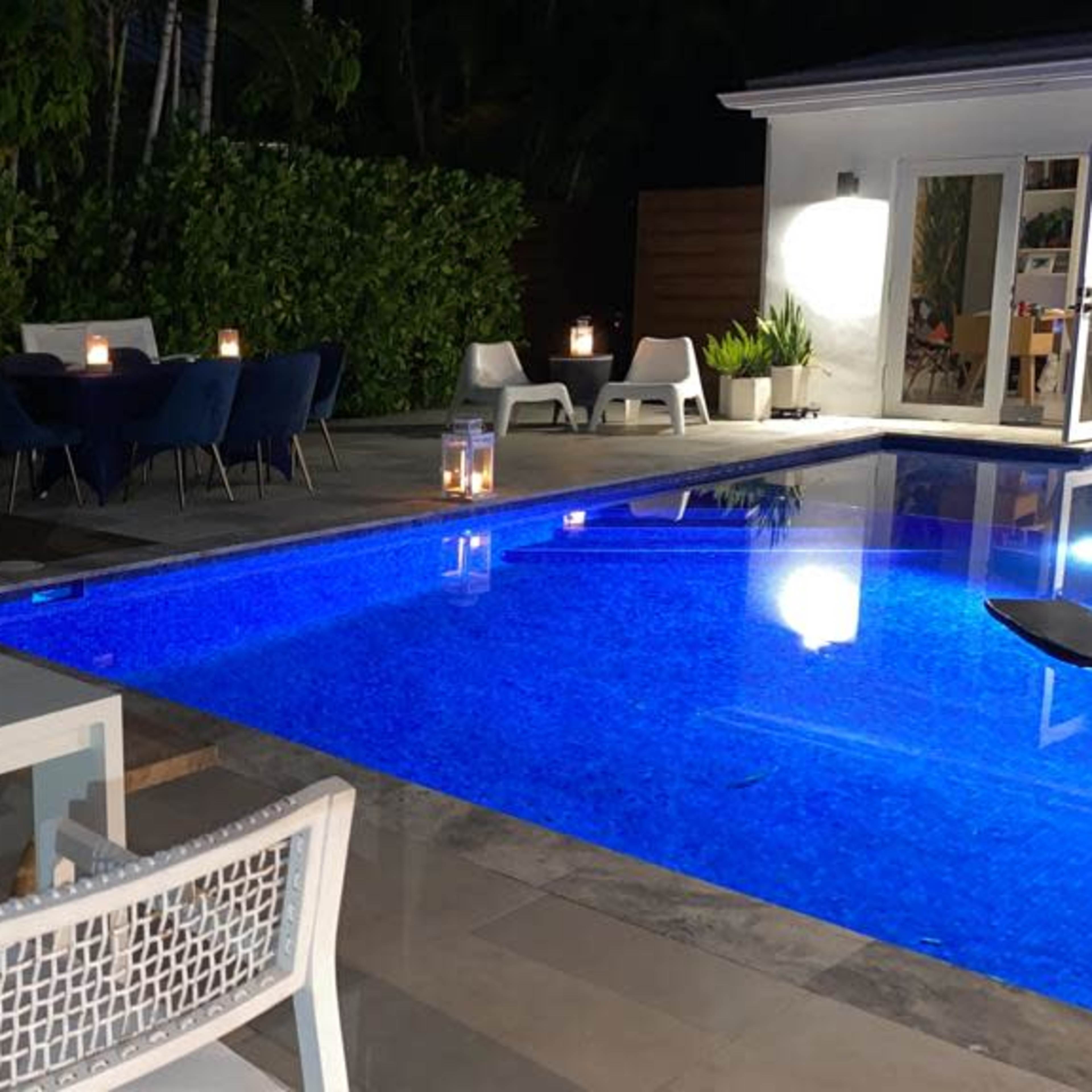 The scene features a well-lit outdoor patio with a dining table and chairs beside a swimming pool surrounded by greenery.
