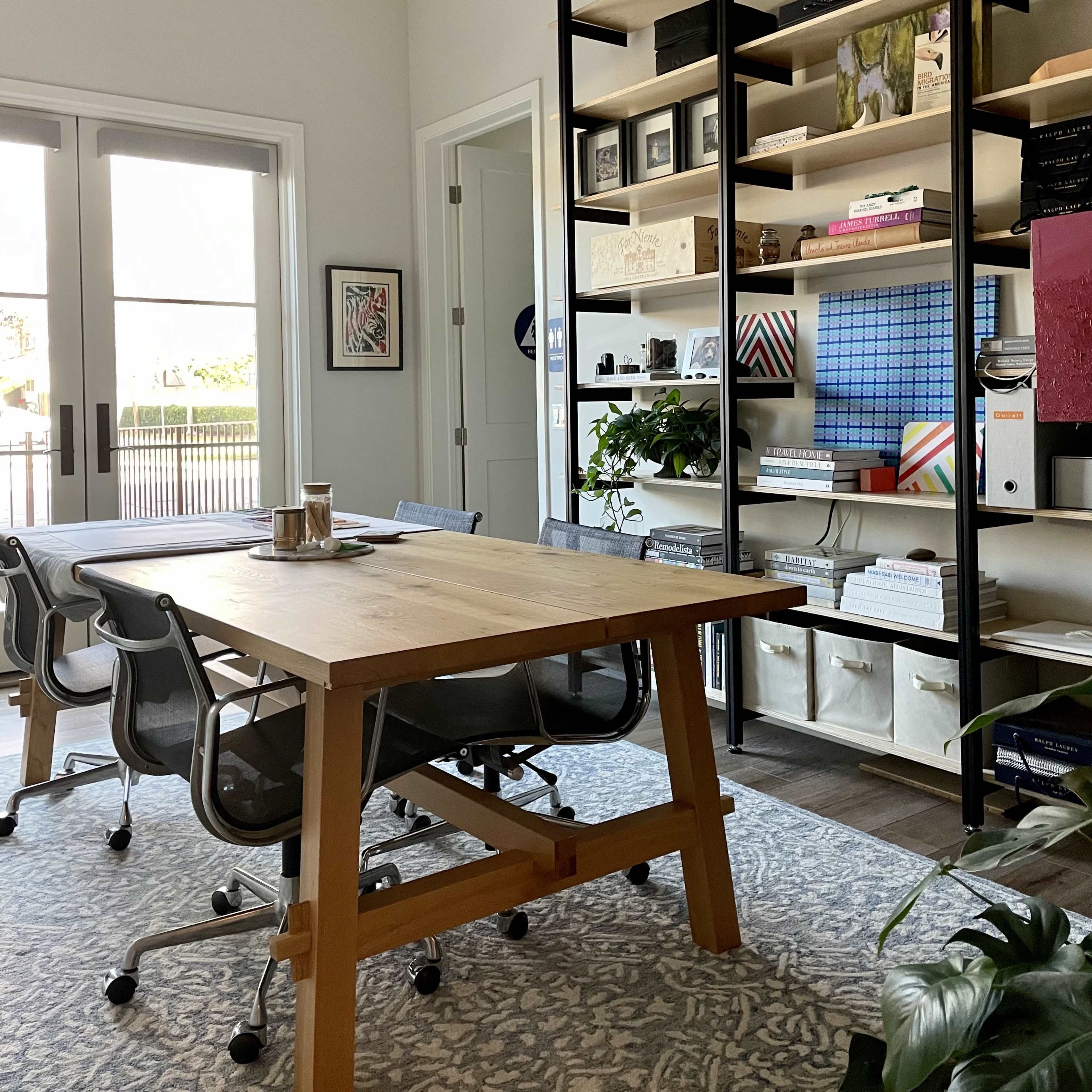 A well-lit office features a wooden table surrounded by chairs, with a shelving unit filled with books and decorative items against the wall.