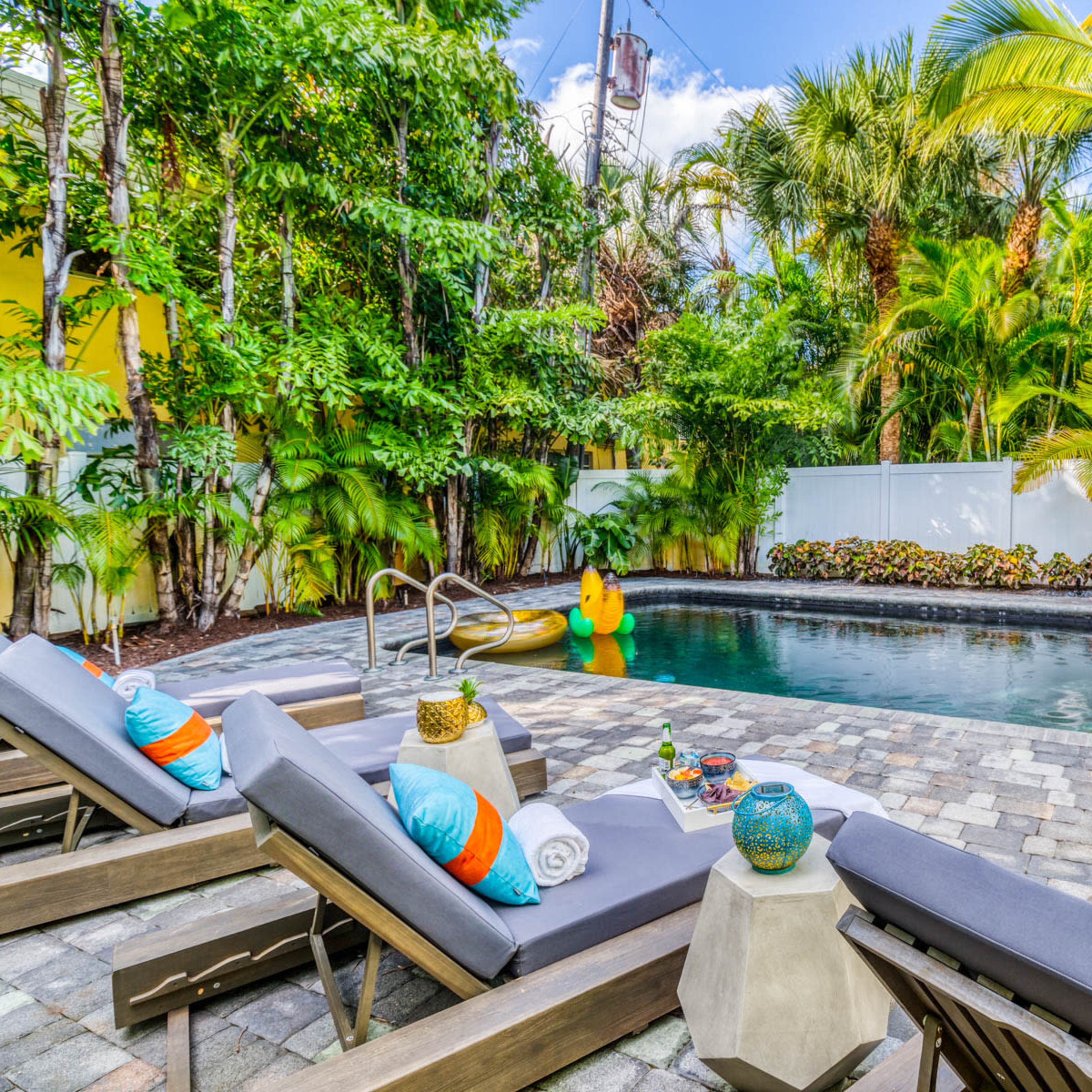 A backyard pool surrounded by lounge chairs, tropical plants, and inflatable pool toys.