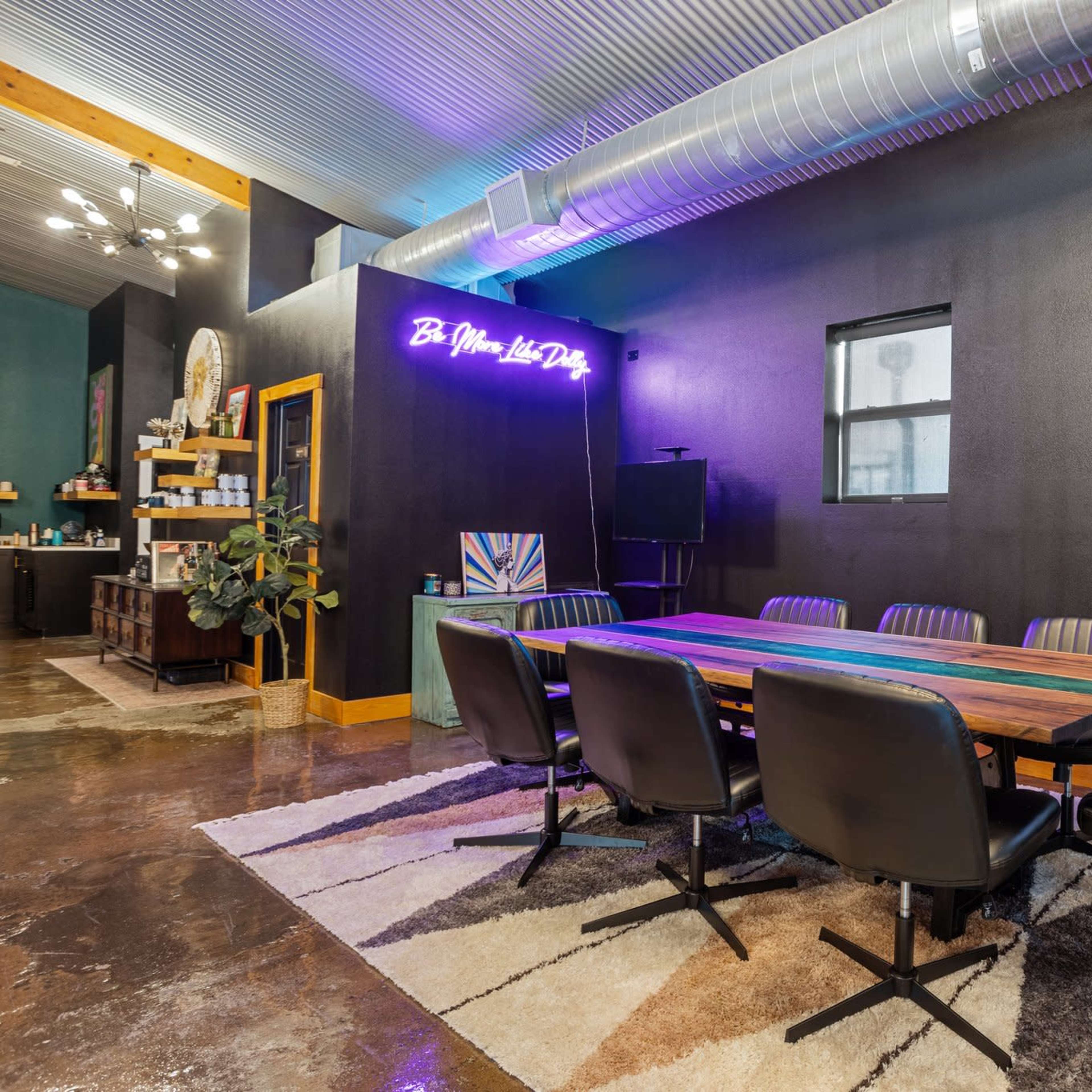 A modern conference room features a large wooden table surrounded by black chairs, with a neon sign on the wall and large windows allowing natural light.