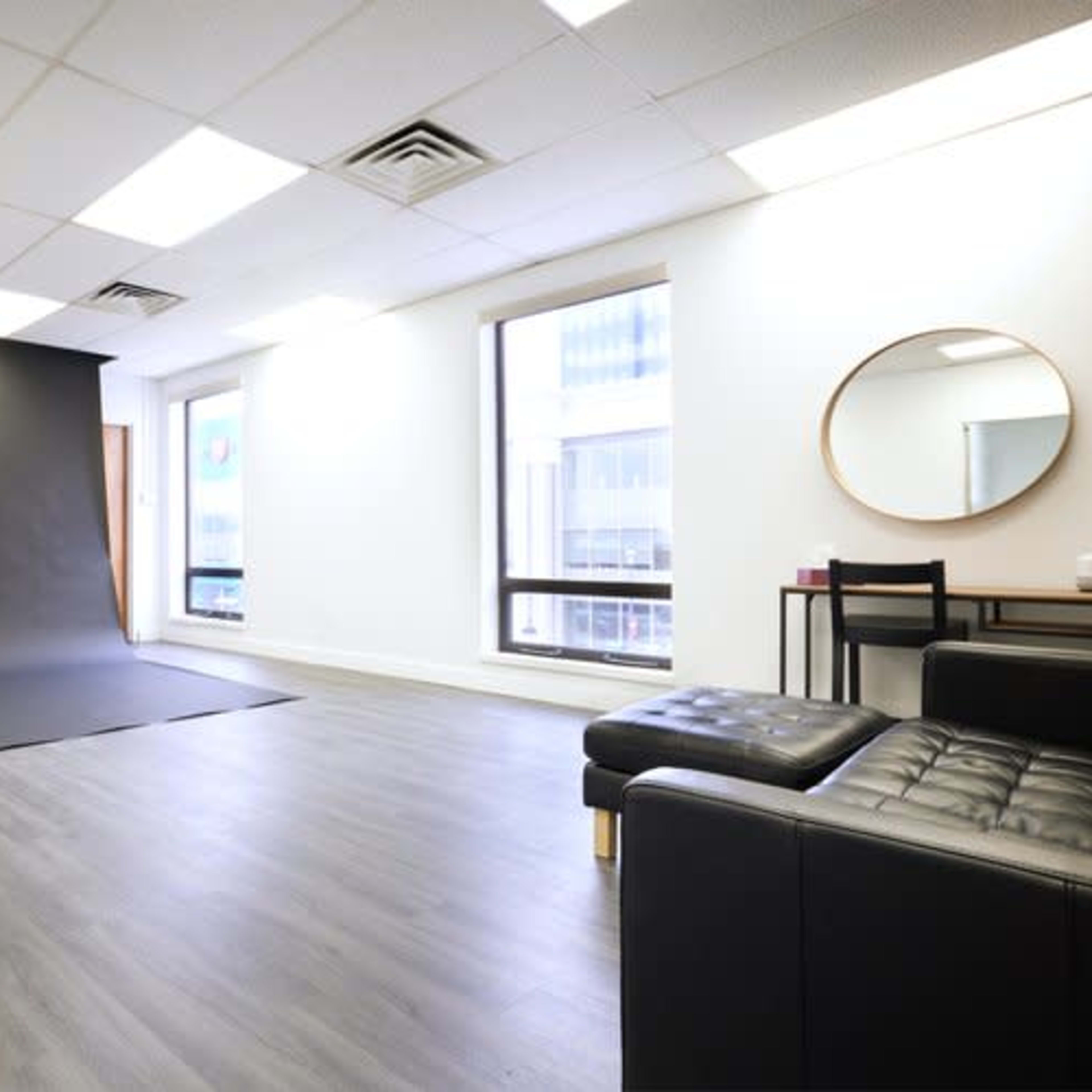 A photography studio with a black backdrop, a light stand, and a modern seating area, featuring large windows that let in natural light.