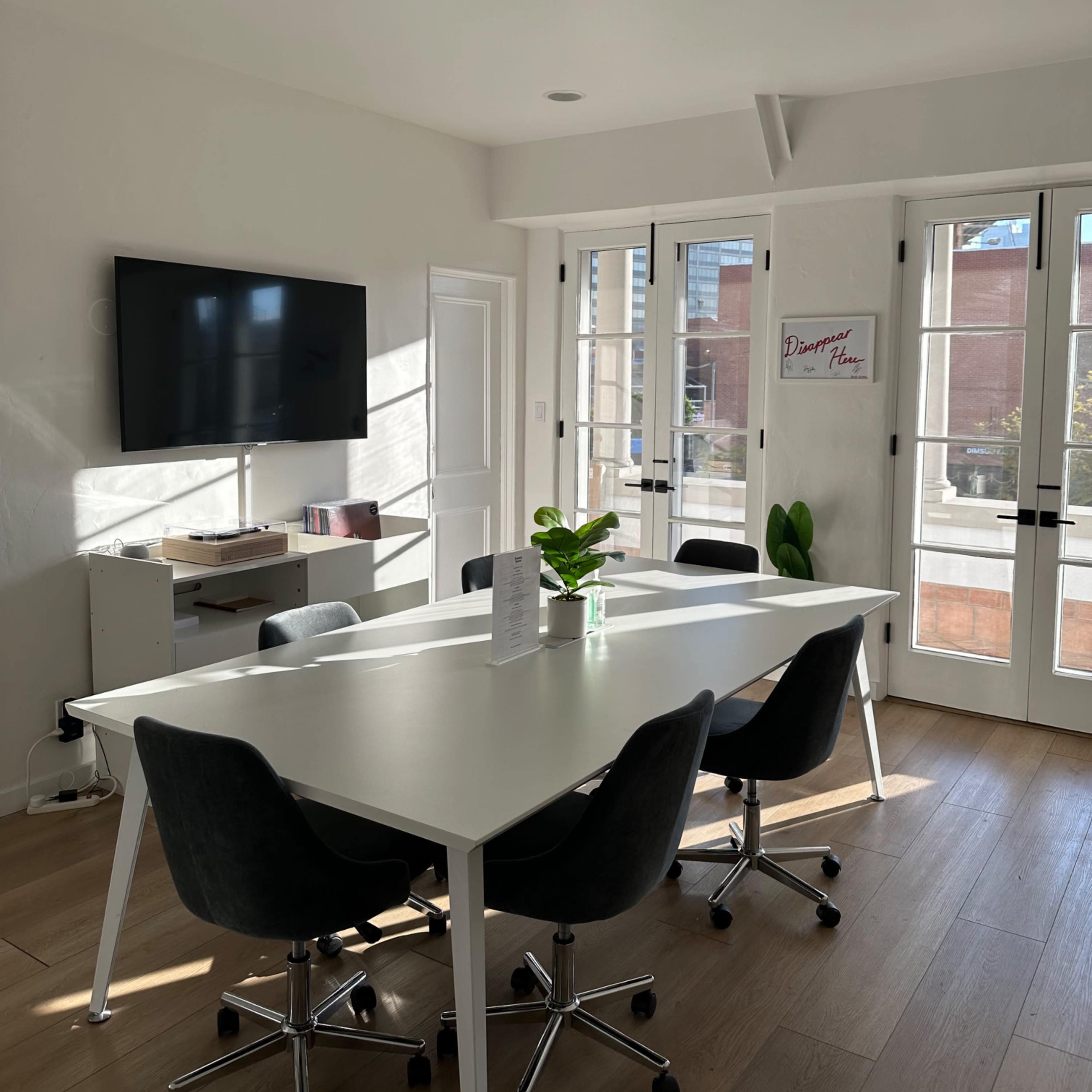 A modern conference room features a long white table surrounded by black rolling chairs, with a large TV mounted on the wall and windows providing natural light.
