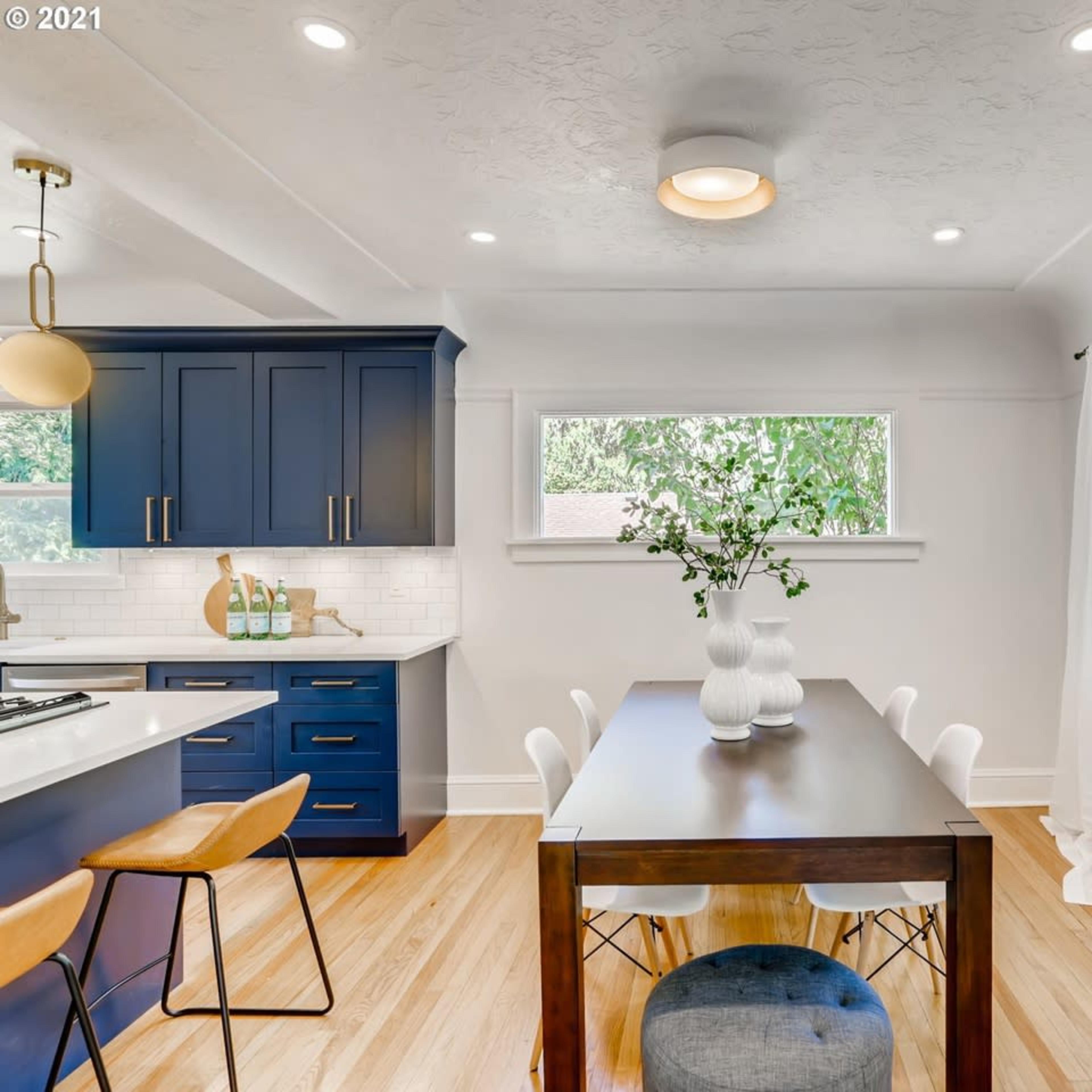 A modern kitchen and dining area features blue cabinetry, a wooden dining table, and large windows allowing natural light.