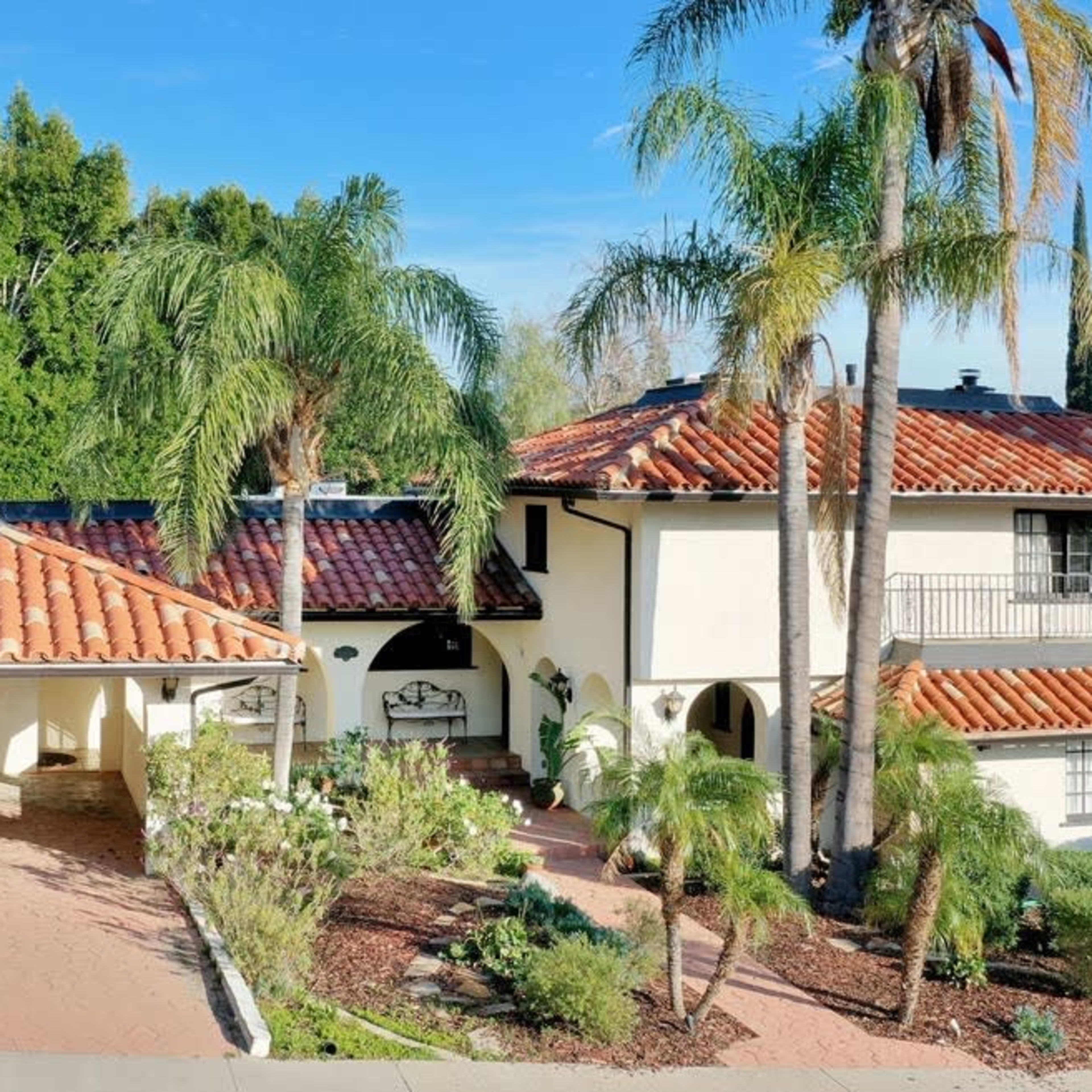 A two-story house with a terracotta roof surrounded by palm trees and landscaped gardens.