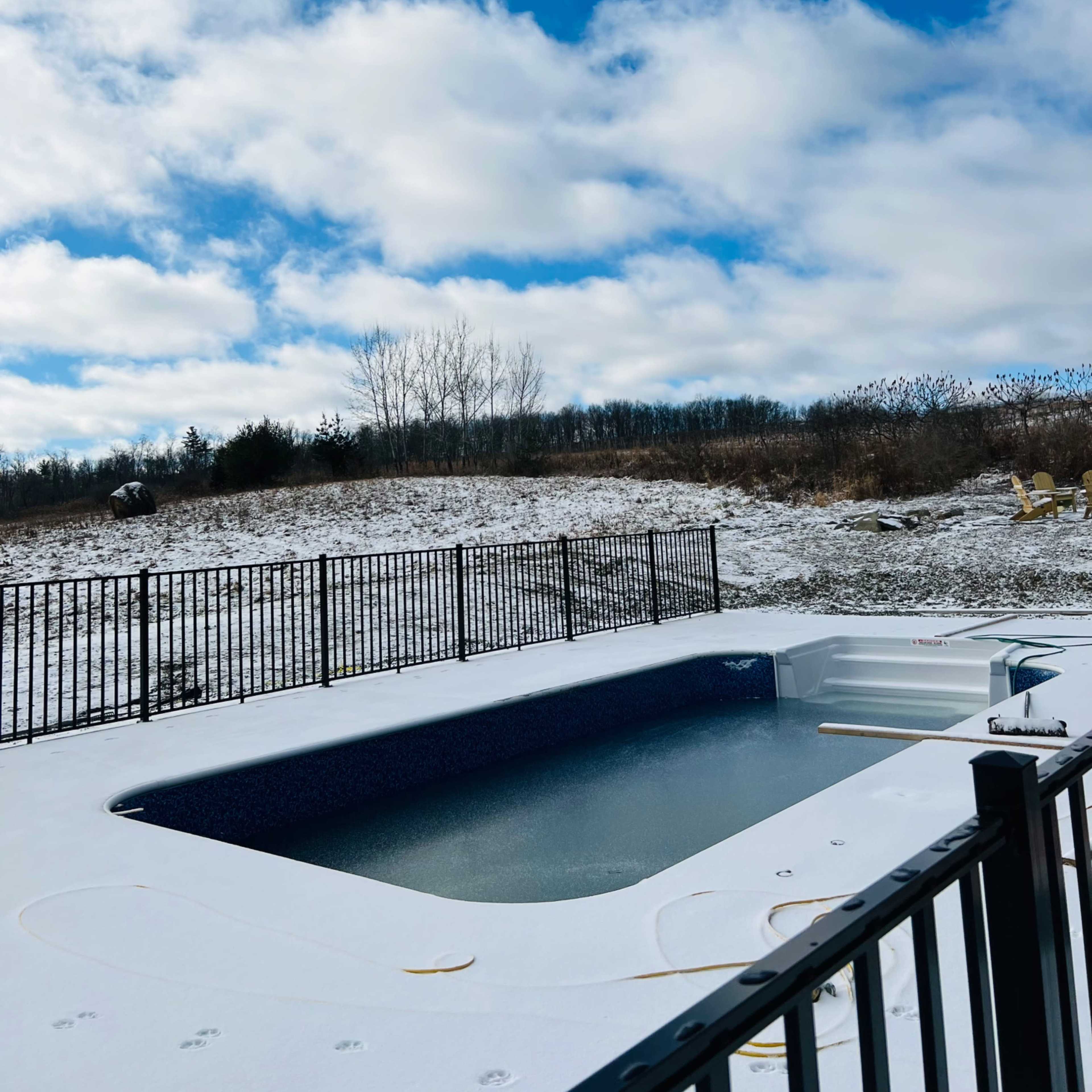 A snow-covered pool area is surrounded by a black fence under a partly cloudy sky.