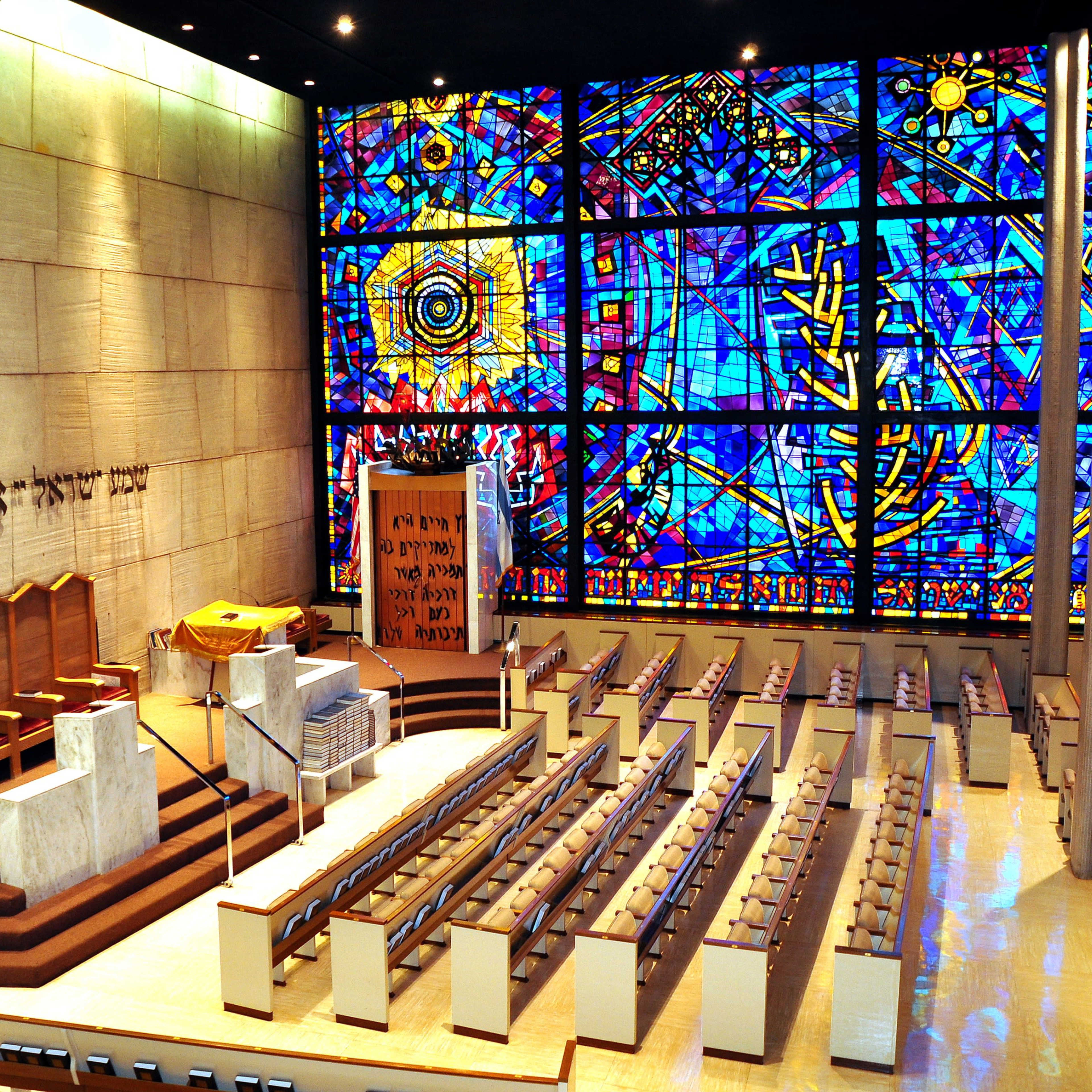 A spacious synagogue interior featuring a large stained glass window, a raised platform with seating at the front, and rows of wooden benches for worshippers.