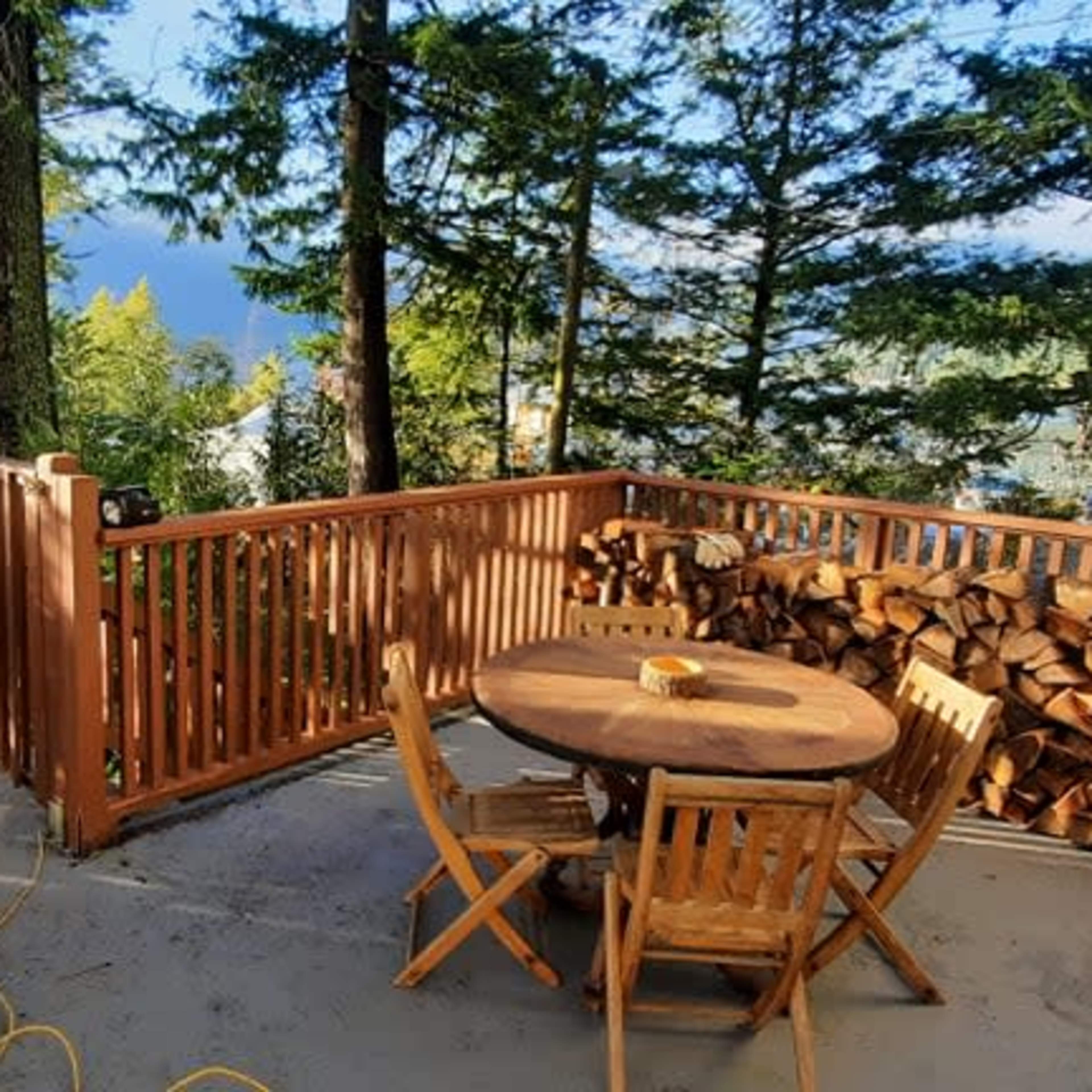 A wooden deck with a round table and chairs overlooks a forested area and stacked firewood.