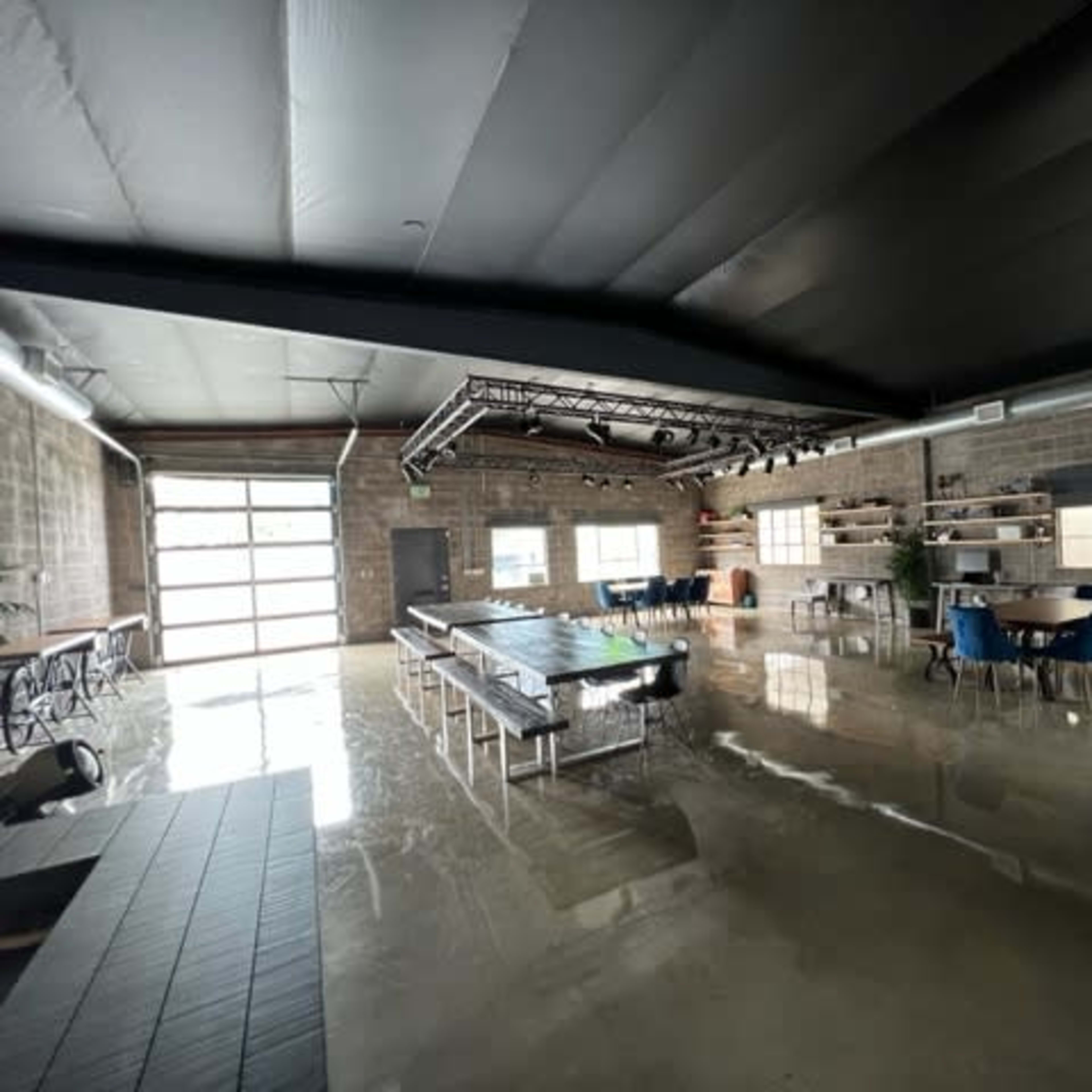 A spacious, industrial-style room with polished concrete floors, featuring a long central table, several dining tables with blue chairs, and stage lighting overhead.